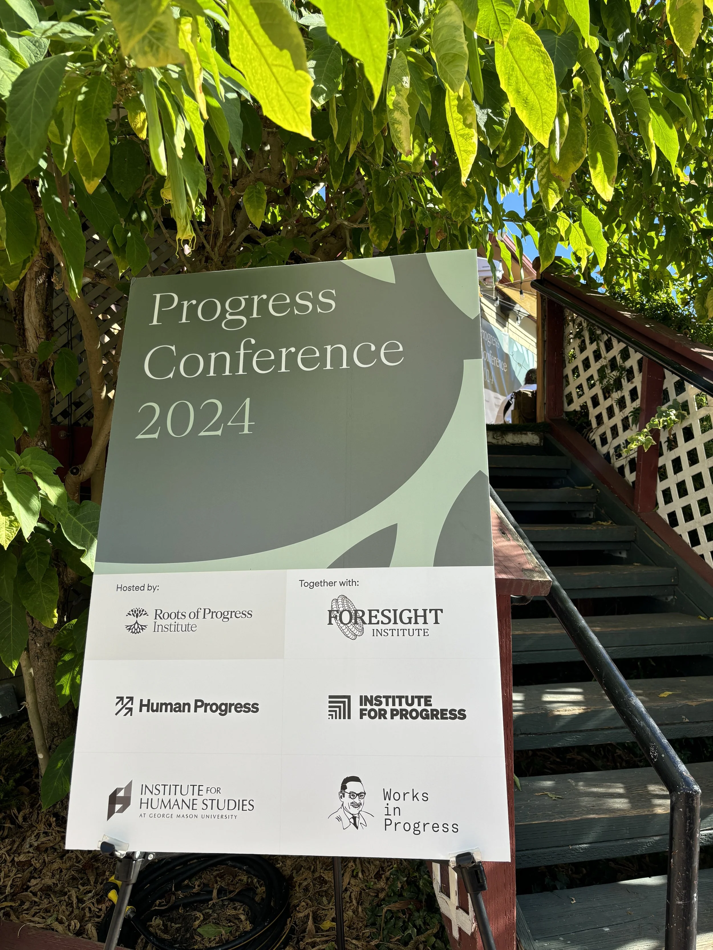 Sign for the Progress Conference 2024, hosted by Roots of Progress Institute and Foresight Institute, with logos of Human Progress, Institute for Progress, Institute for Humane Studies, and Works in Progress, placed outside near a staircase and green