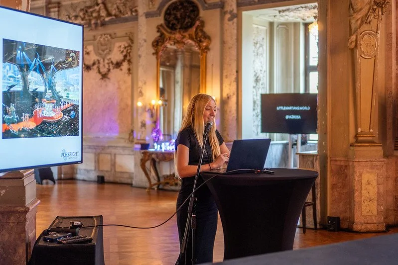A woman with long blonde hair standing at a podium with a laptop, speaking into a microphone in an ornate historical room with high ceilings, chandelier, decorative mirrors, and large windows. A large screen displaying an image of a space-themed digi