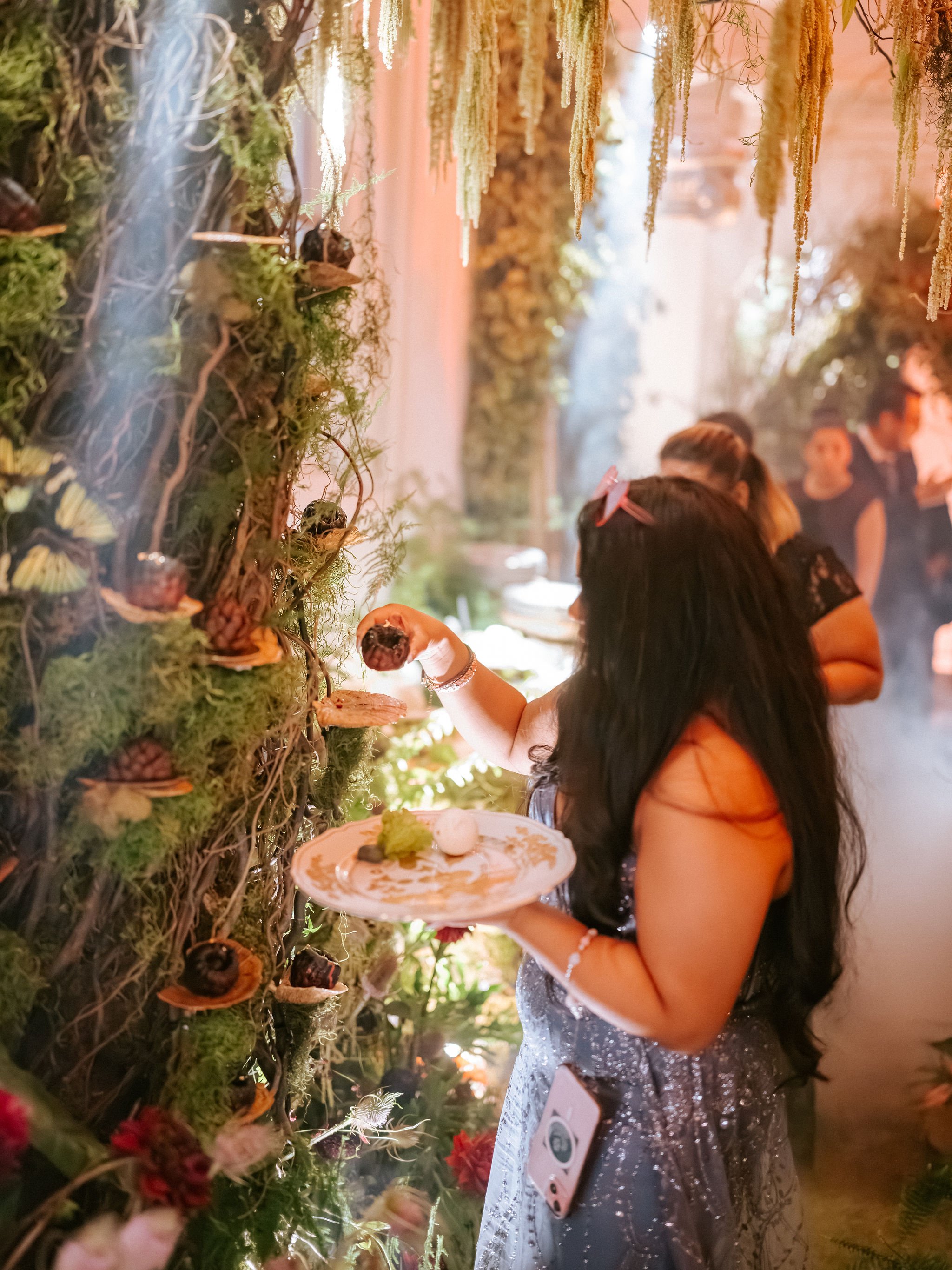 A woman is at a sushi or food station, holding a plate with food and using chopsticks to pick up a piece of sushi. She has long dark hair, is wearing a sparkly dress and has a phone clipped to her dress. The station has a decorative, moss-covered wal