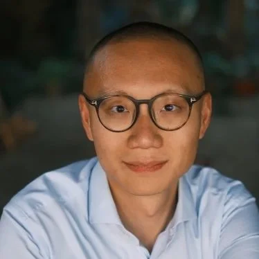 Close-up of a person with glasses wearing a white shirt, smiling against a blurred outdoor background.