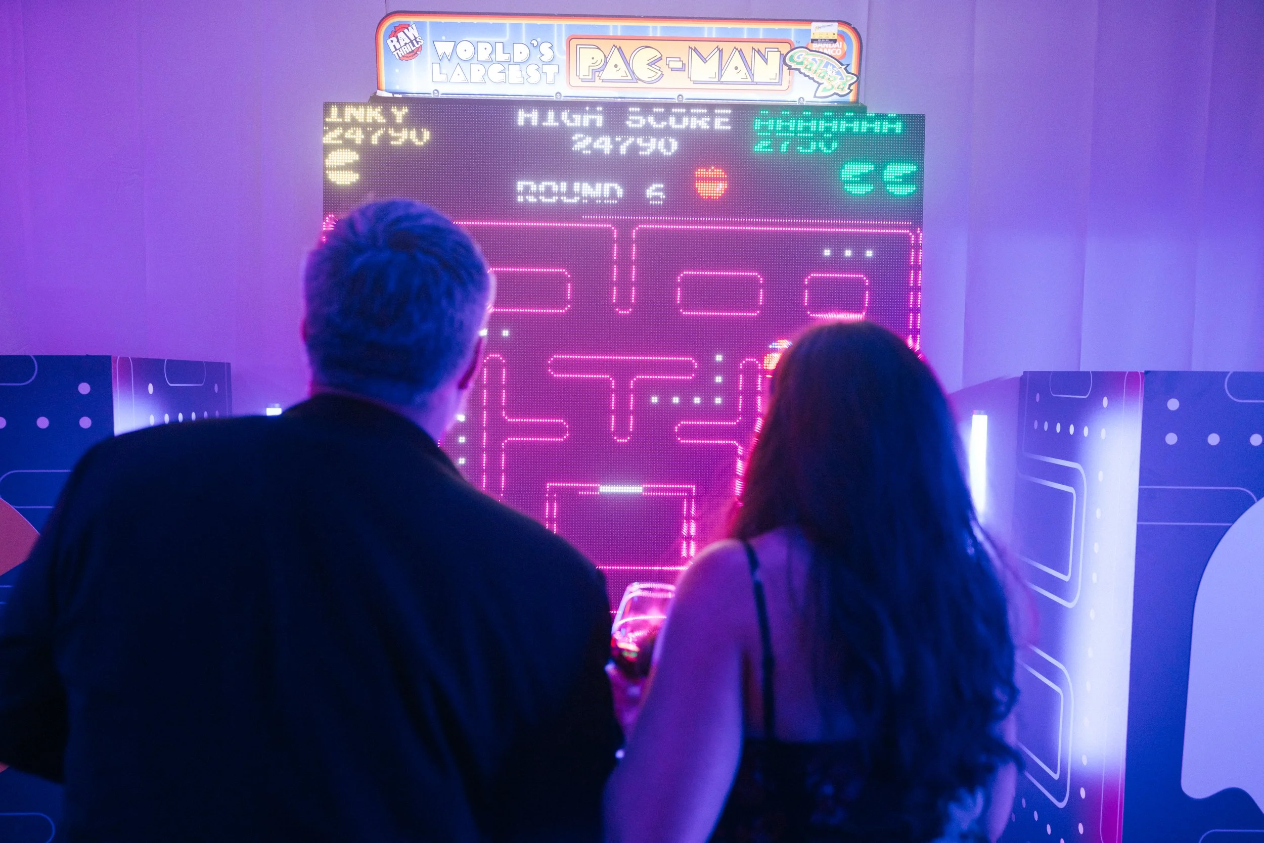 Two people playing a Pac-Man arcade game in a neon-lit setting, viewed from behind. The game screen shows a maze with dots, and the score and round number are visible at the top.