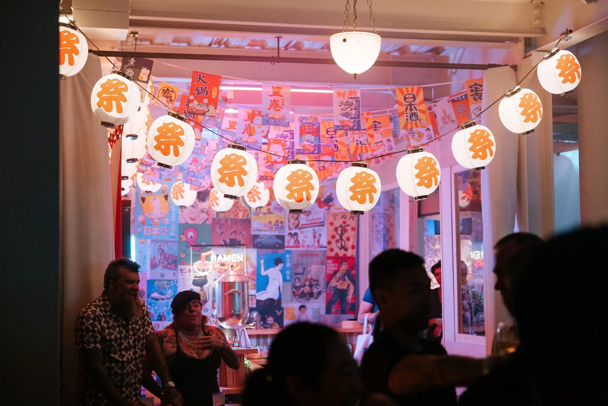Interior of a Japanese-themed restaurant decorated with paper lanterns featuring kanji characters, colorful banners, and posters on the walls. There are people socializing inside.