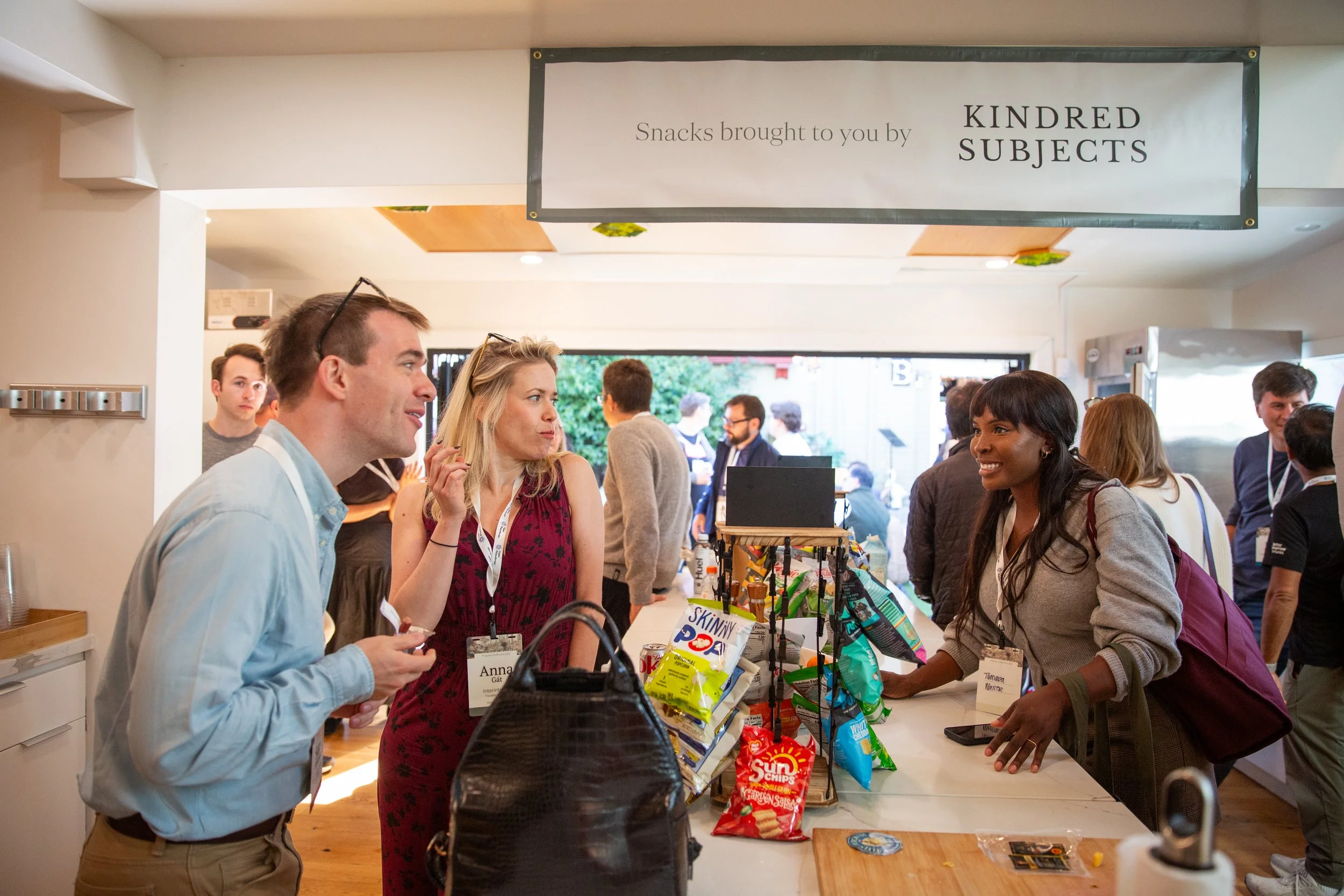 People shopping at a snack stand with a sign overhead that reads 'Snacks brought to you by KINDRED SUBJECTS', featuring various snack bags and a cashier.