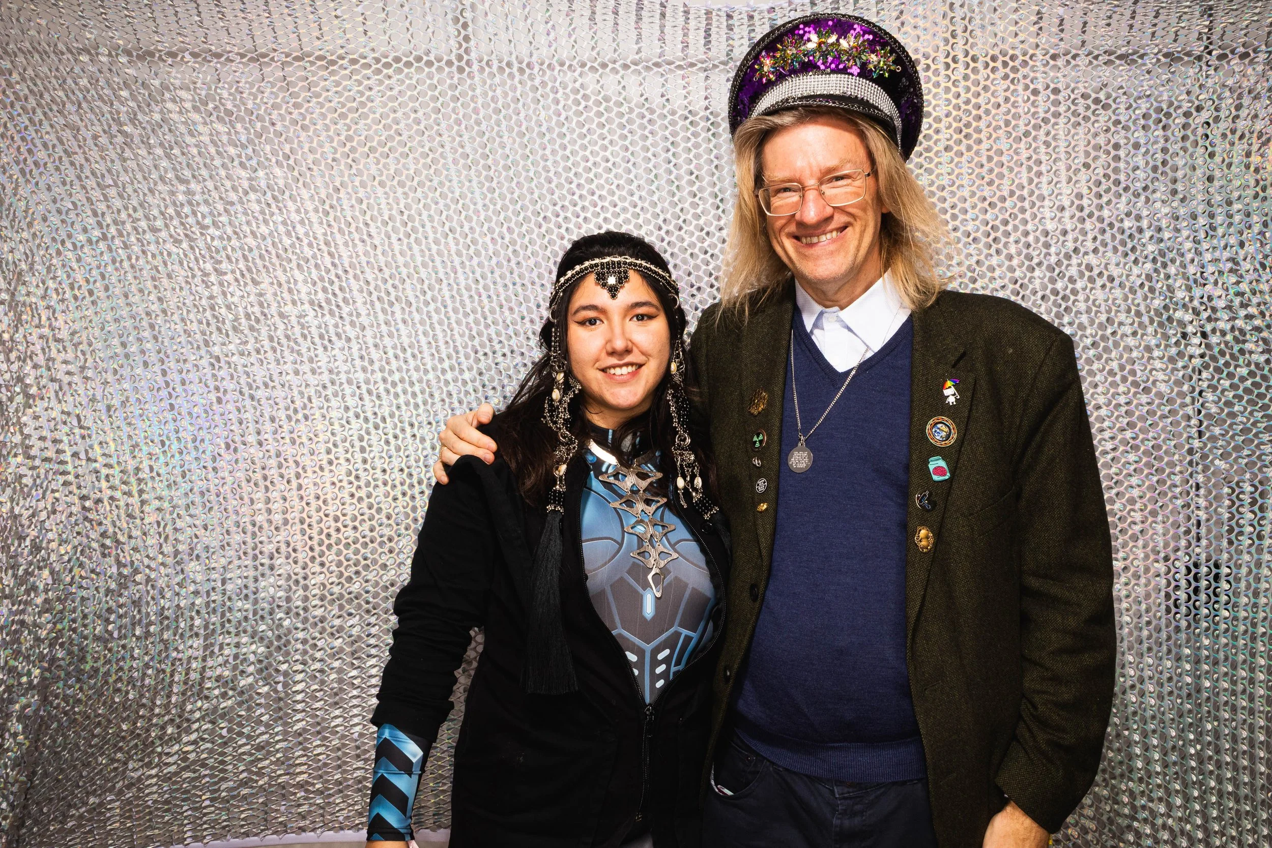 Two people smiling and posing together, with one person wearing a futuristic and sci-fi themed outfit and the other person dressed in a casual blazer with pins and a hat, standing against a textured, metallic silver background.