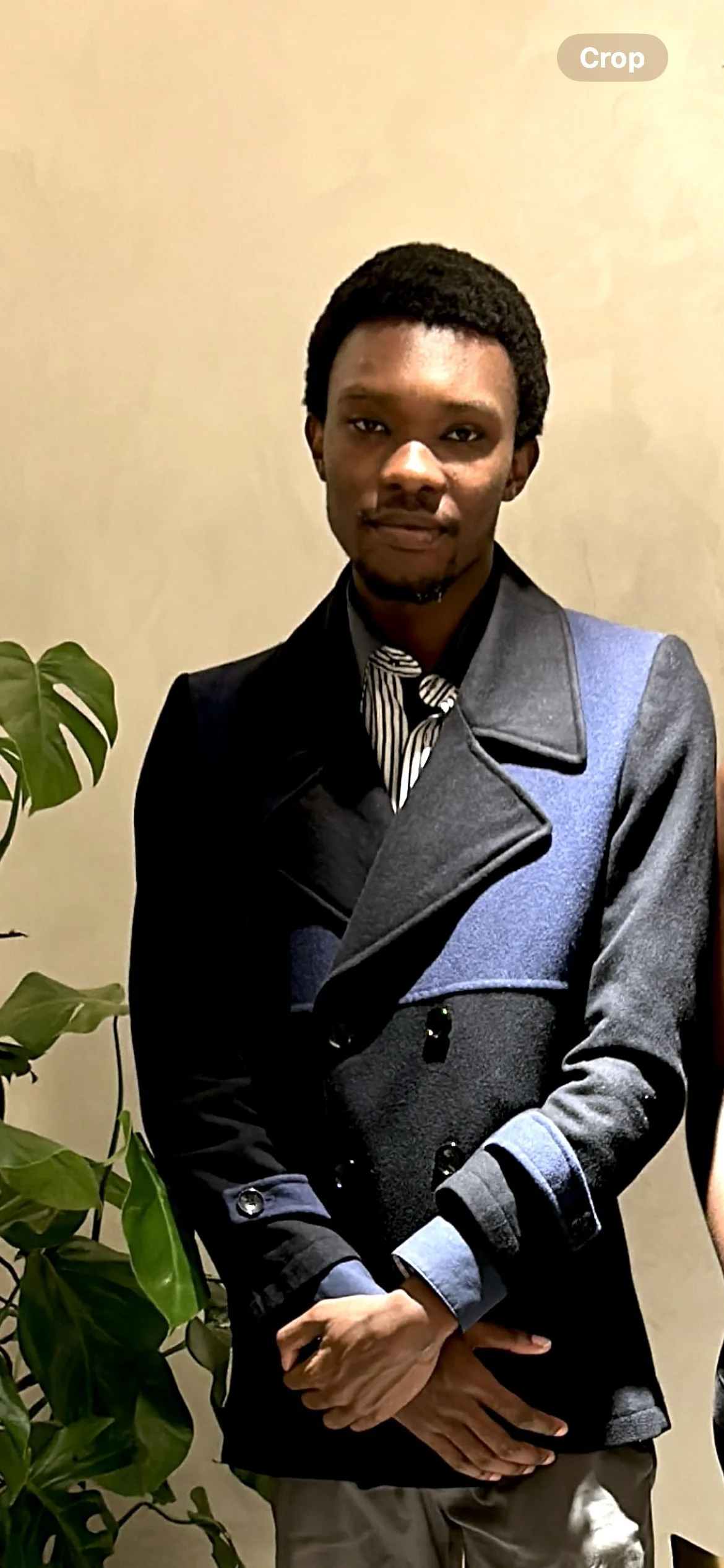 A man with dark hair and a beard wearing a stylish gray and black coat over a striped shirt, standing next to a green potted plant against a beige wall.