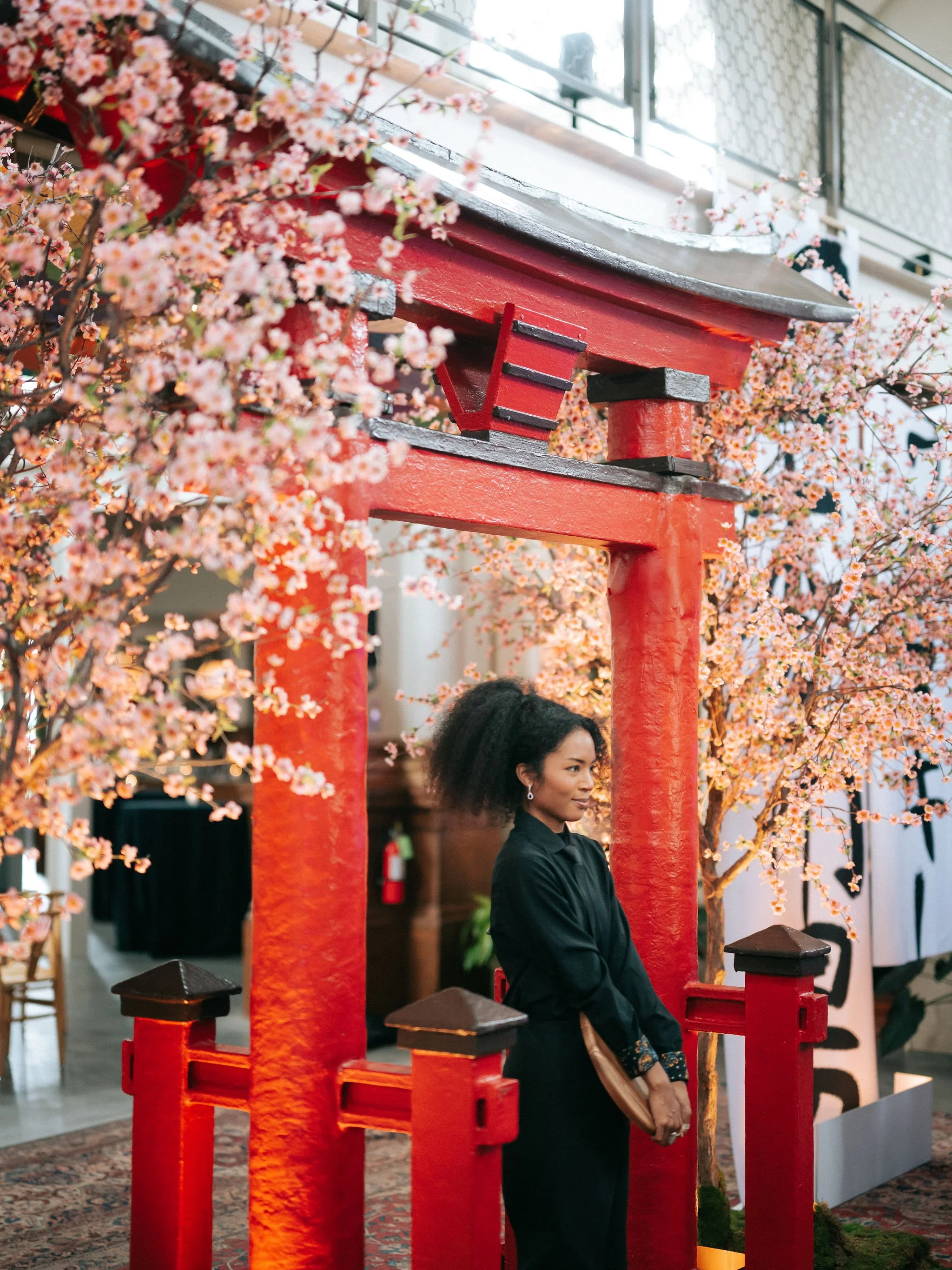 A woman with dark curly hair stands inside a red torii gate surrounded by pink cherry blossoms, indoors.
