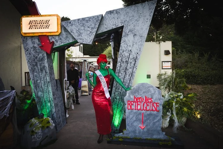 A woman dressed as the Grinch with green face paint and a red dress, wearing a sash that reads 'Mistress of Mirth,' standing next to a gravestone with the words 'here lies Betelgeuse' and a red arrow pointing down. She is inside a decorative archway labeled 'Betelgeuse.' The scene appears to be a Halloween or themed event with decorations and plants around.