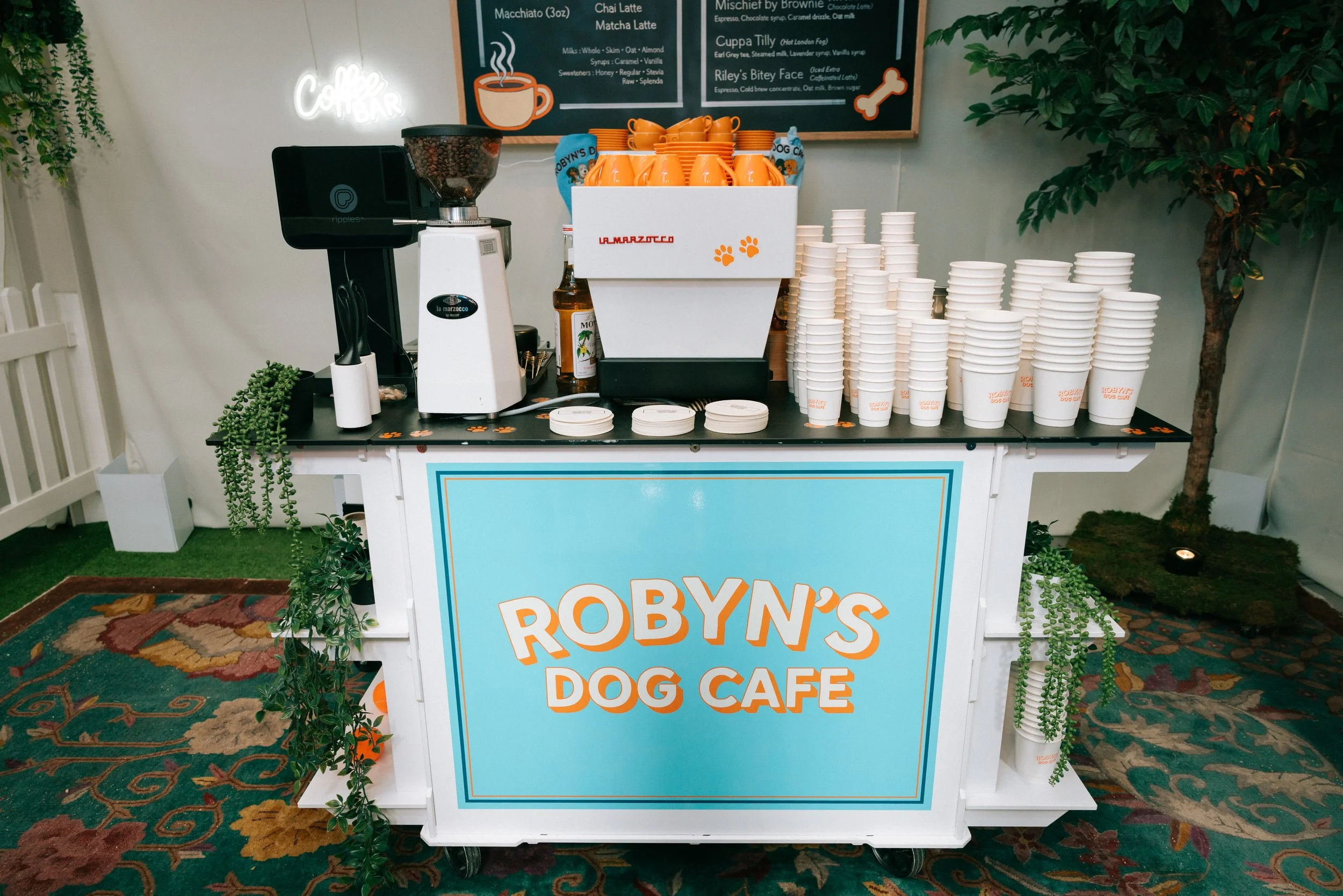 A coffee and drink station at Robyn's Dog Cafe, with a large sign on the front, stacks of cups, a coffee grinder, a blender, and a tray of orange cups with paw prints, surrounded by plants and a patterned carpet.