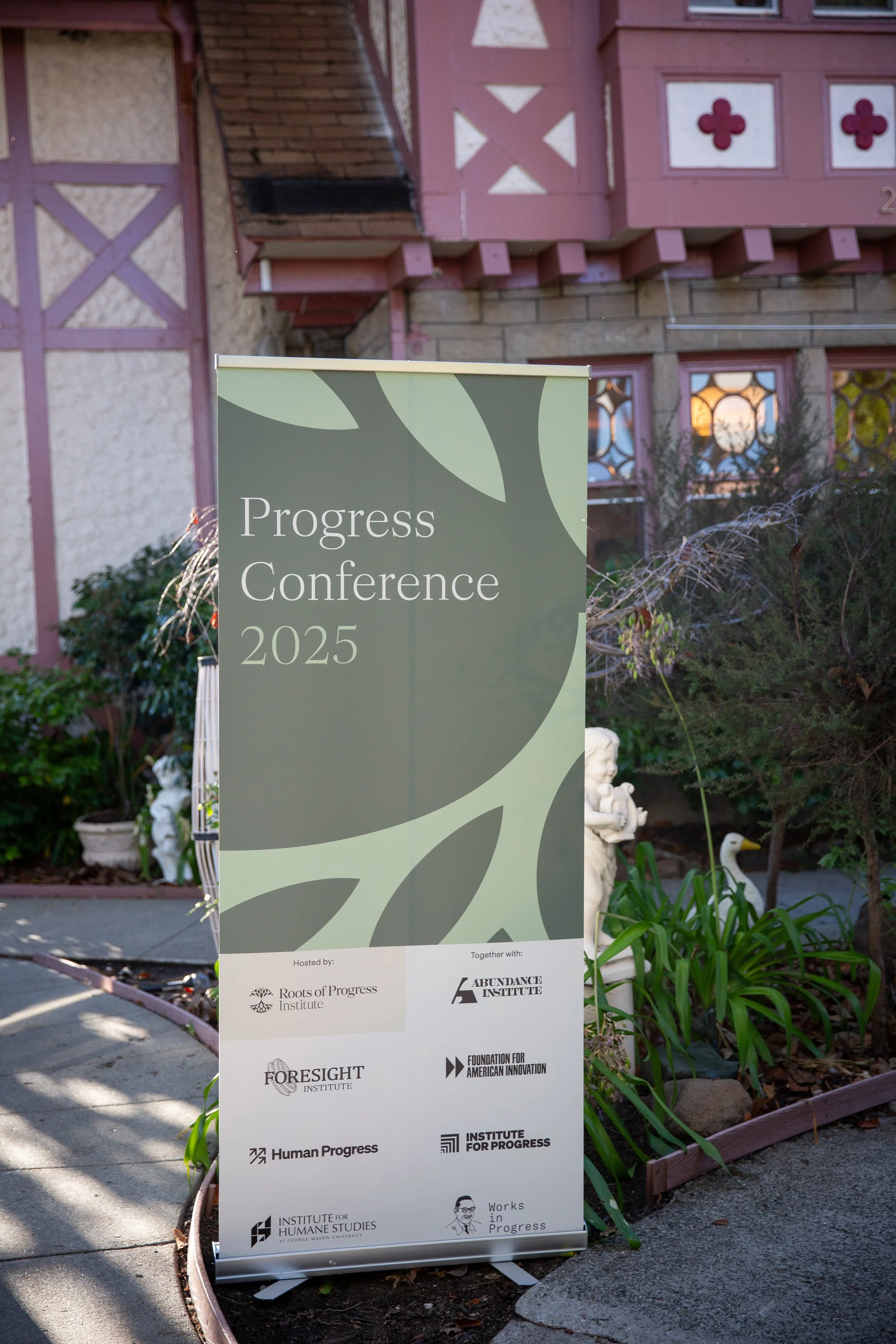 A vertical sign in front of a house reads 'Progress Conference 2025' and displays logos of various organizations including Roots of Progress Institute, Abundance Institute, Foresight Institute, Foundation for American Innovation, Human Progress, Inst