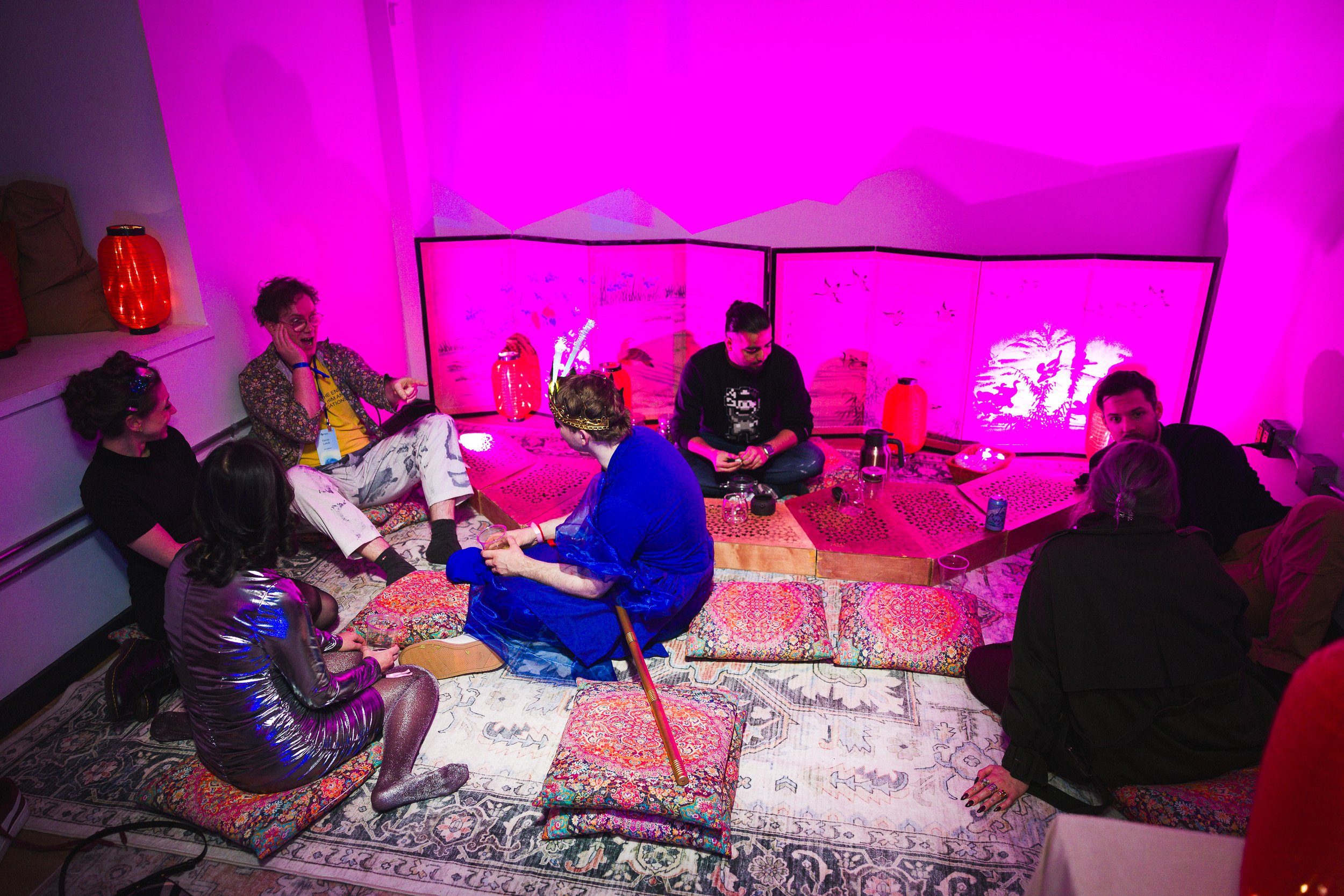 A group of people sitting on a colorful floor surrounded by cushions and pillows, illuminated by pink and red lighting, with a decorative folding screen behind them with landscape artwork, some holding drinks and engaging in conversations at a social