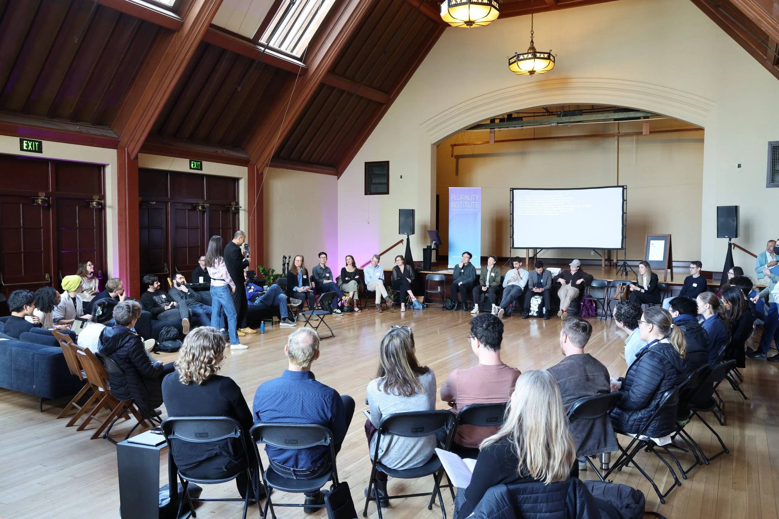 A group of people seated in a circular arrangement in a large, wood-paneled room with high ceilings, attending a presentation or discussion. A person stands in the middle speaking into a microphone, while other participants listen. A large screen at 