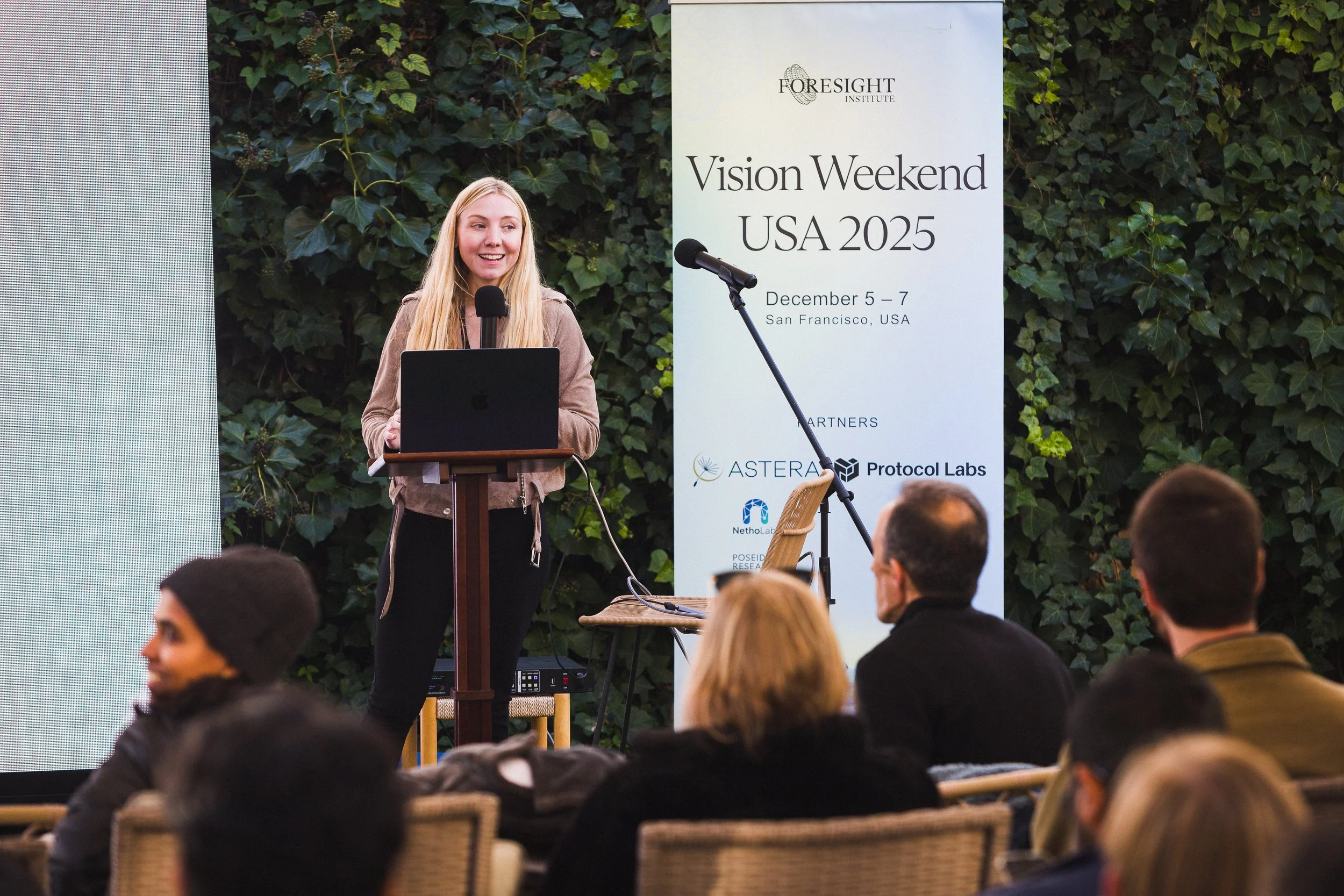 A young woman with blonde hair giving a presentation at the Vision Weekend USA 2025 event, standing behind a podium with a laptop, in front of a green leafy background and a large event banner, with several attendees seated in front.