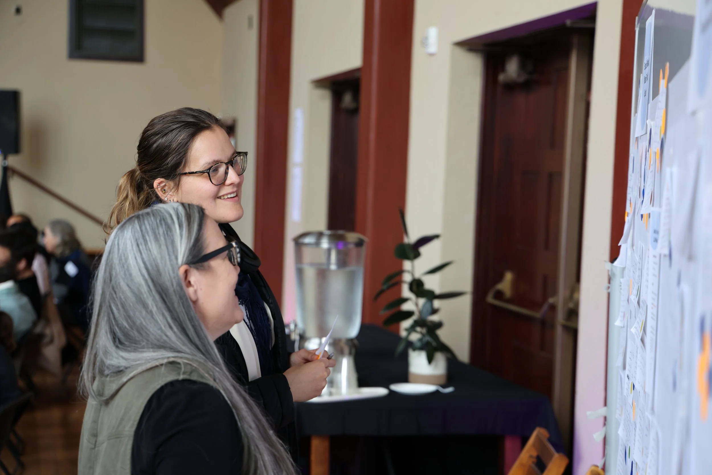 Two women with glasses looking at a bulletin board filled with papers and notes in a conference or event setting.
