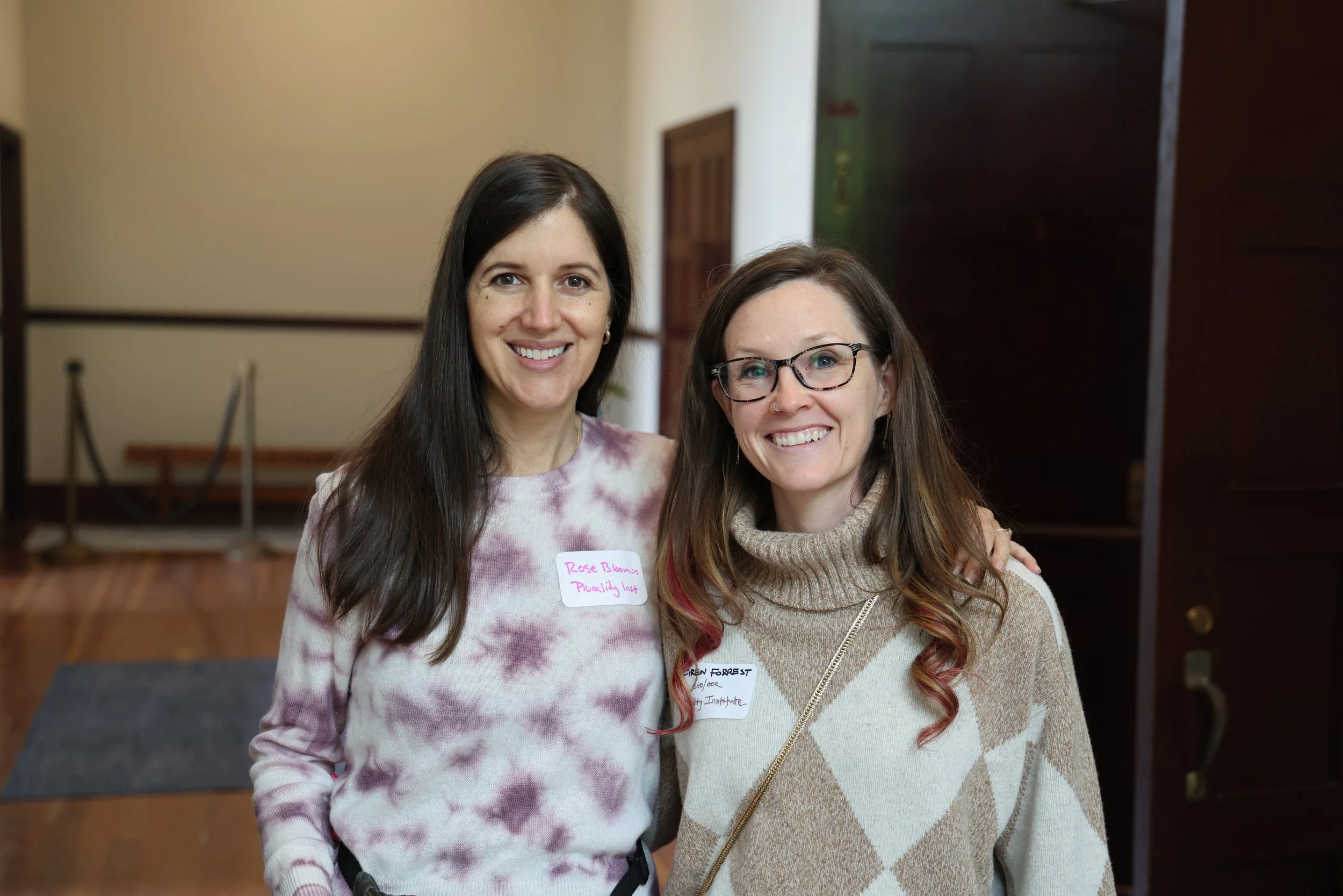 Two women standing indoors, smiling at the camera. One woman has long dark hair, wearing a tie-dye shirt, and the other has shoulder-length brown hair, glasses, and a turtleneck sweater. Both women have name tags.