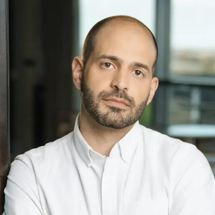 Man with a beard and shaved head, wearing a white button-up shirt, standing indoors near a glass window.