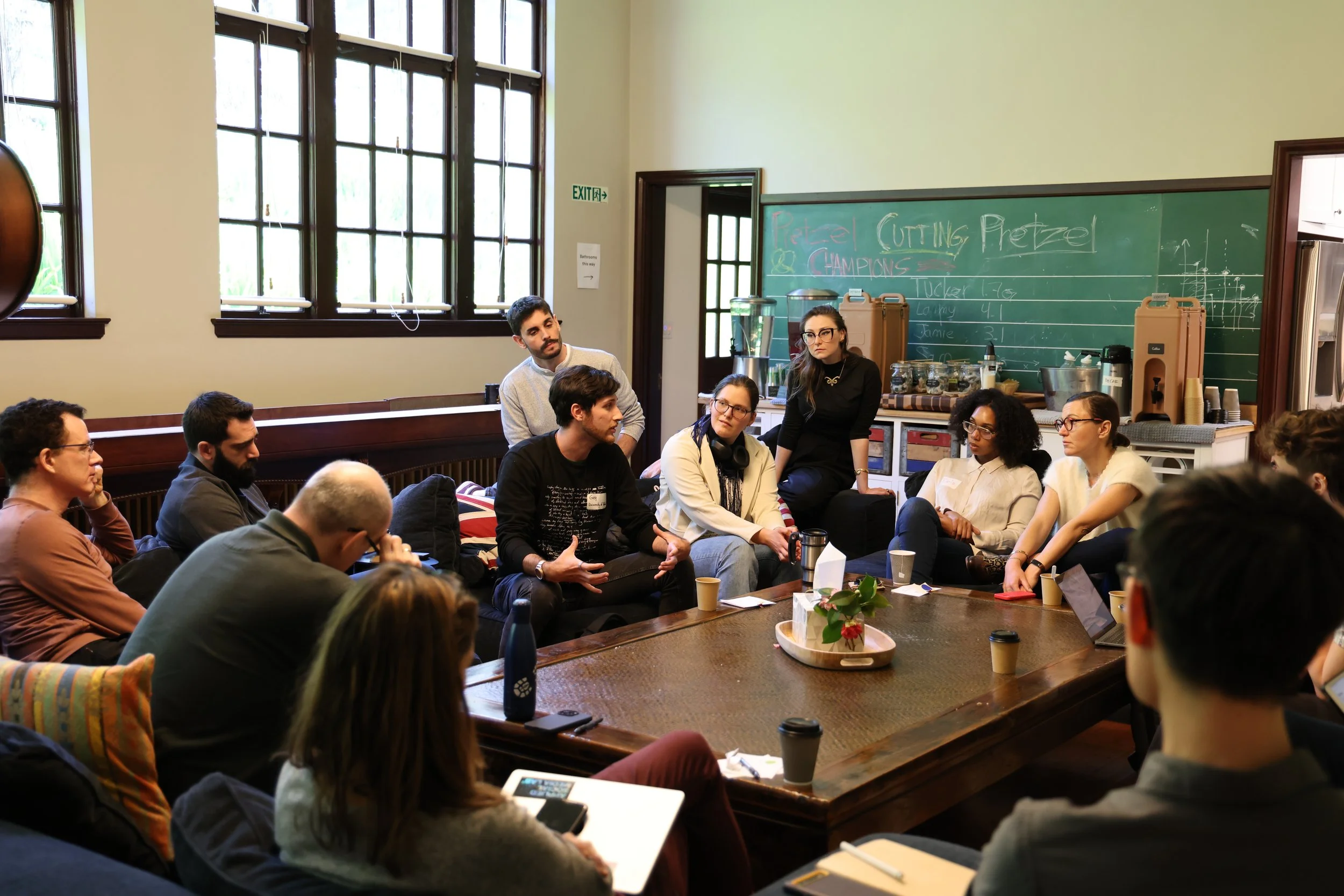 A group of people in a room with large windows, sitting around a long table, engaged in a discussion or meeting, with a chalkboard in the background.