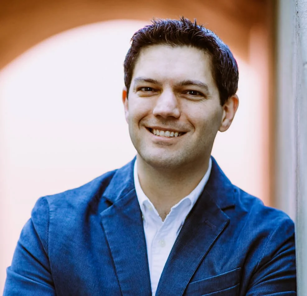 A smiling man in a blue blazer and white shirt standing outdoors near a wall with a blurred background.