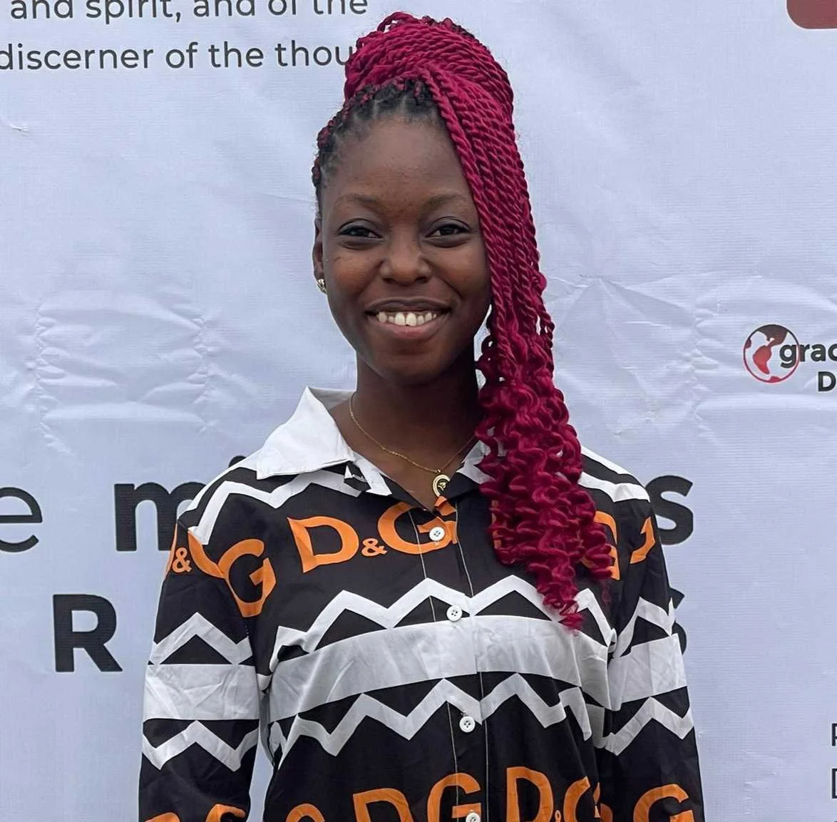 A young woman with dark skin and long red twisted braids smiling, standing in front of a white background with text and a logo.