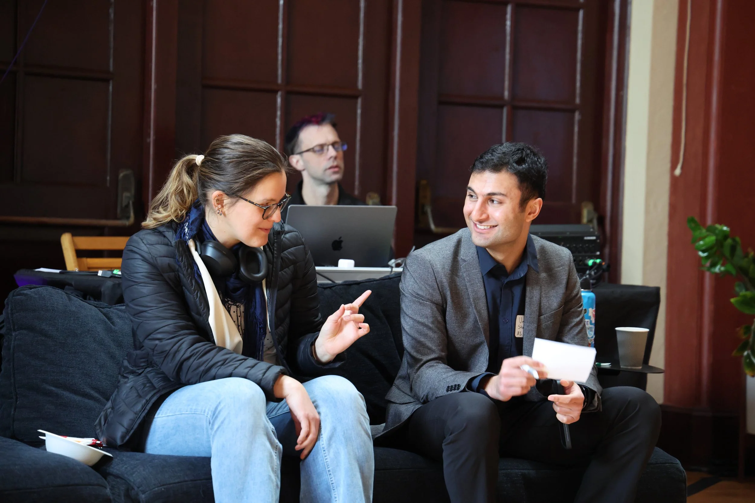 Three people in a meeting room: a woman with glasses, headphones around her neck, and a man in a suit, sitting on a couch and talking, while another man with glasses and a laptop is in the background.