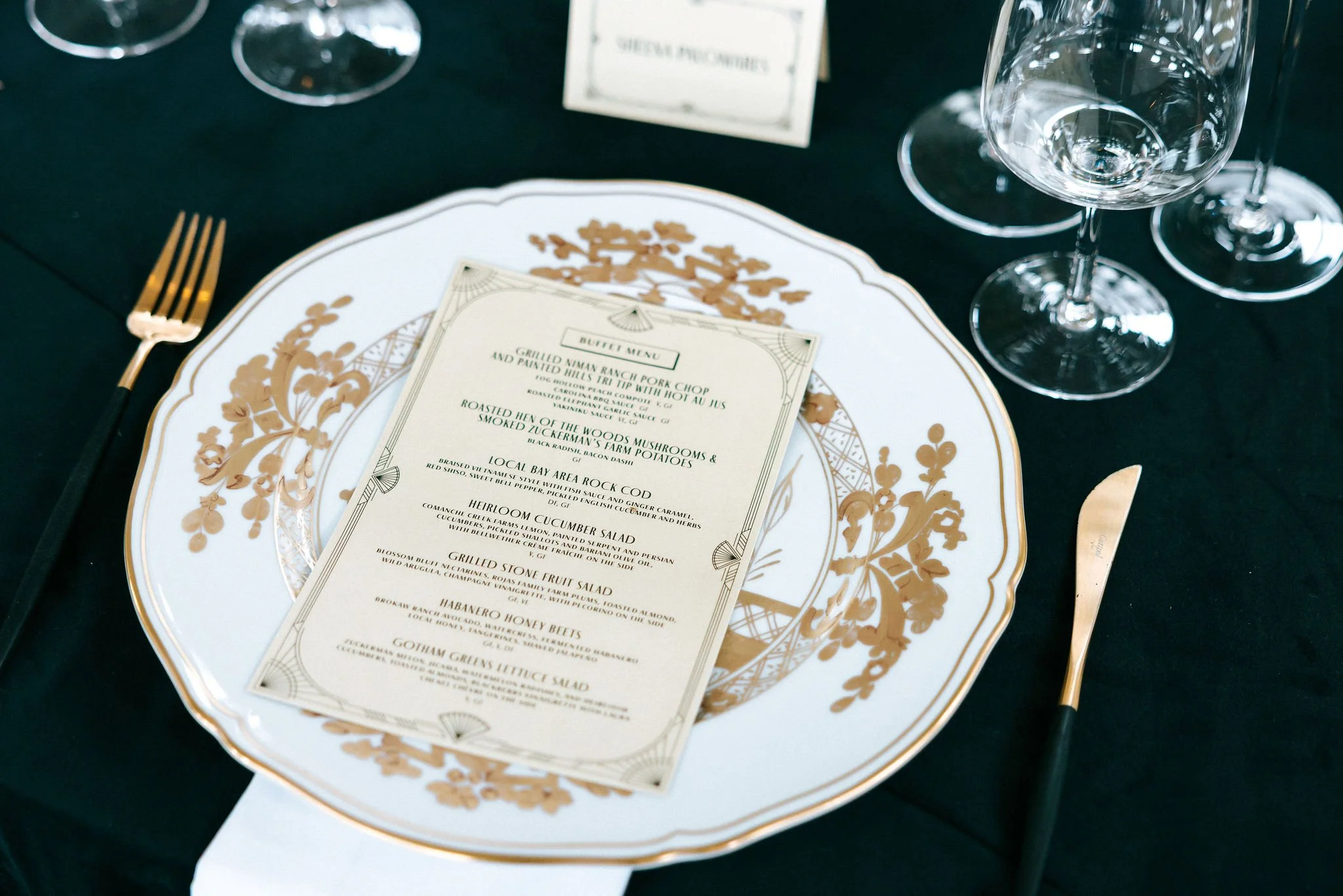 A decorative white and gold plate with a black and gold fork and knife placed on a dark tablecloth, holding a menu with food options and a few empty wine glasses nearby.