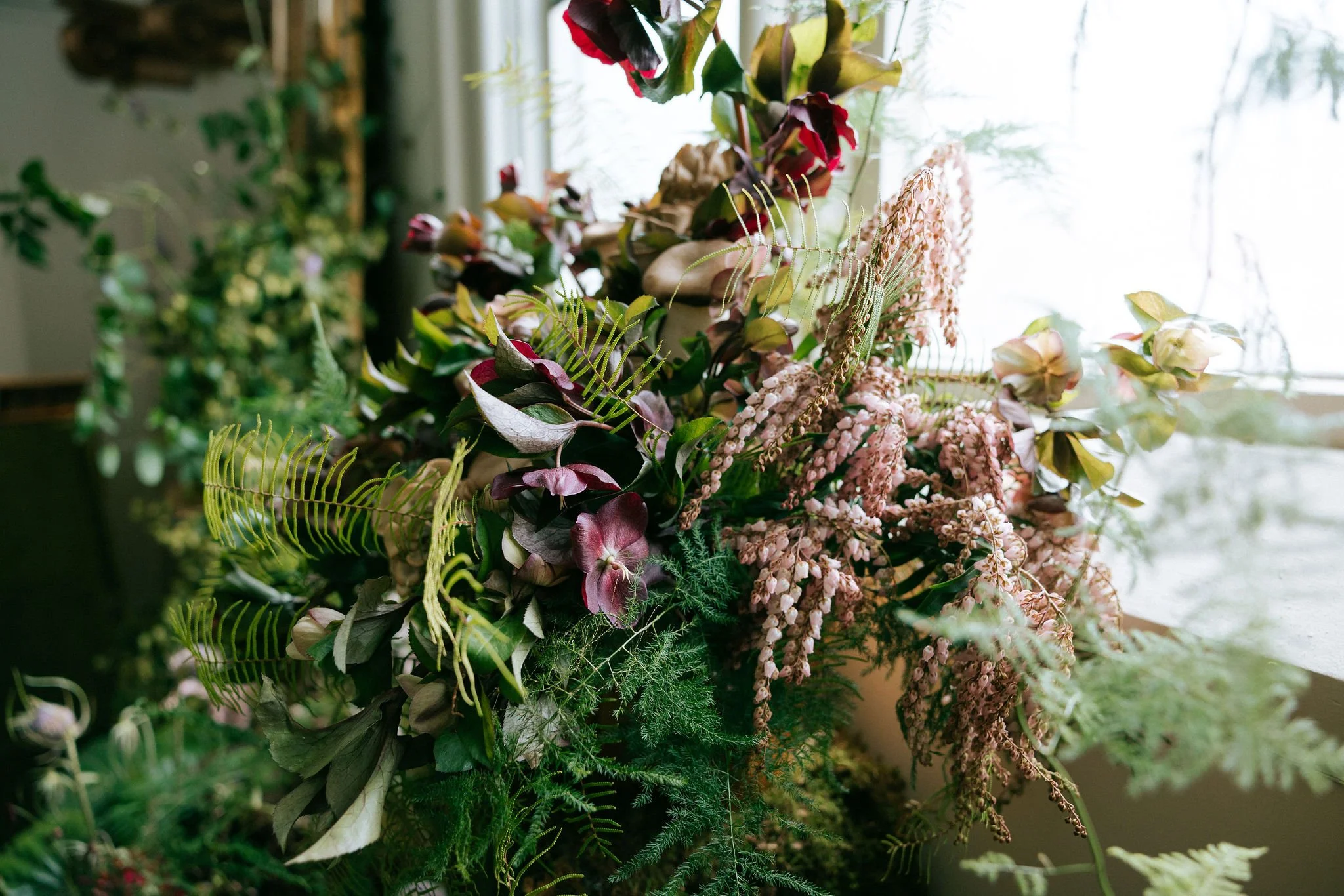 A large bouquet of mixed flowers and greenery near a window, including pink, purple, and cream-colored blooms with various ferns and leaves.