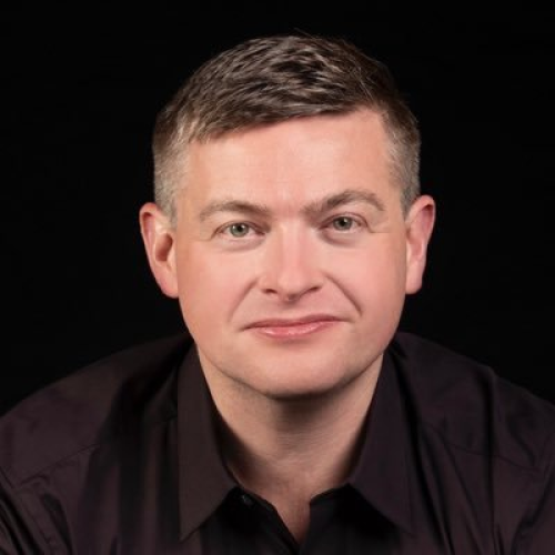 A portrait of a man with short light brown hair, wearing a black shirt, against a black background.