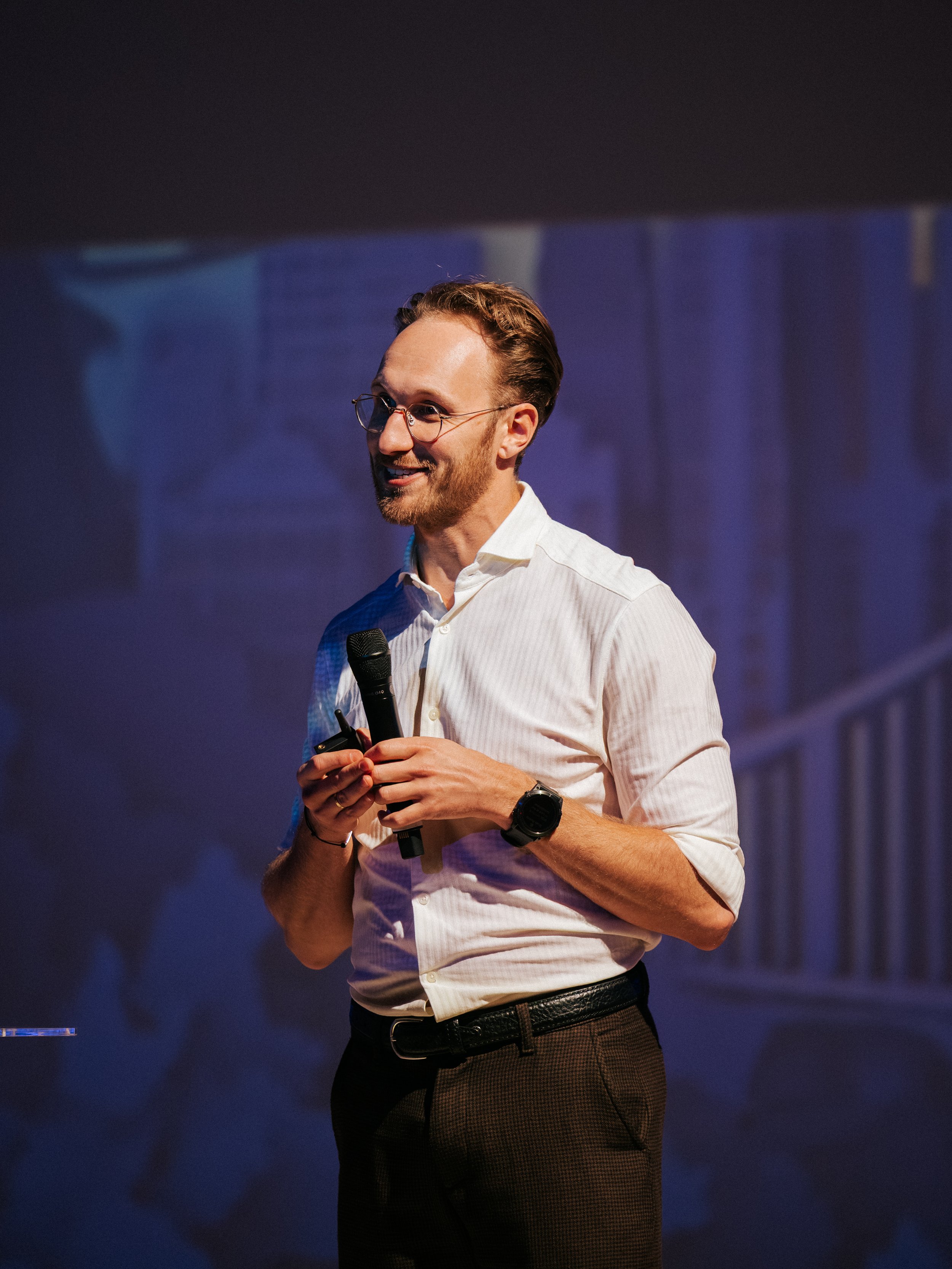 A man with glasses, a beard, and brown hair holding a microphone and a clicker on stage, smiling, wearing a white dress shirt, brown trousers, and a watch, with a dark background.