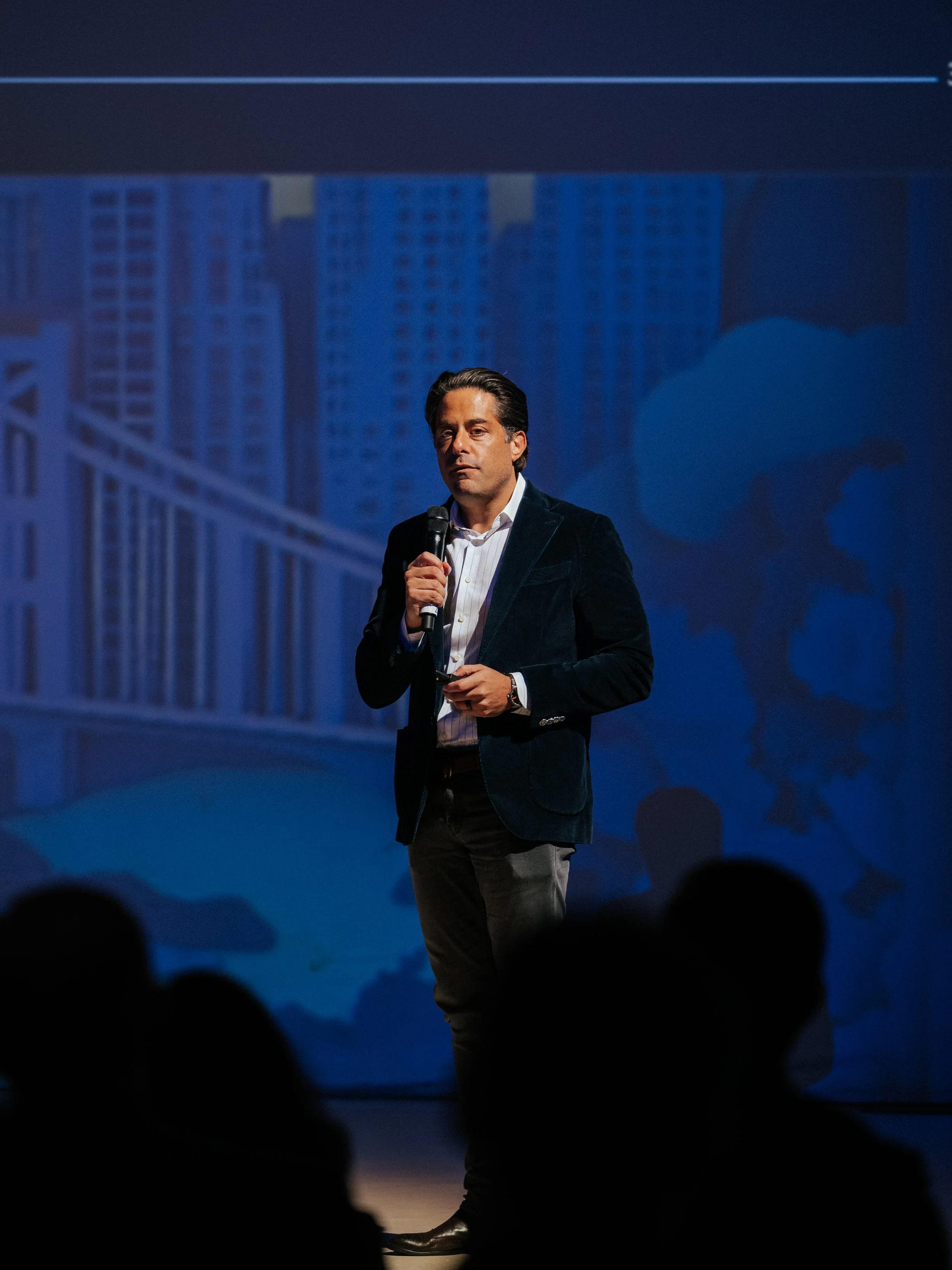 A man in a dark blazer and light shirt holding a microphone on stage with a cityscape background projected behind him.