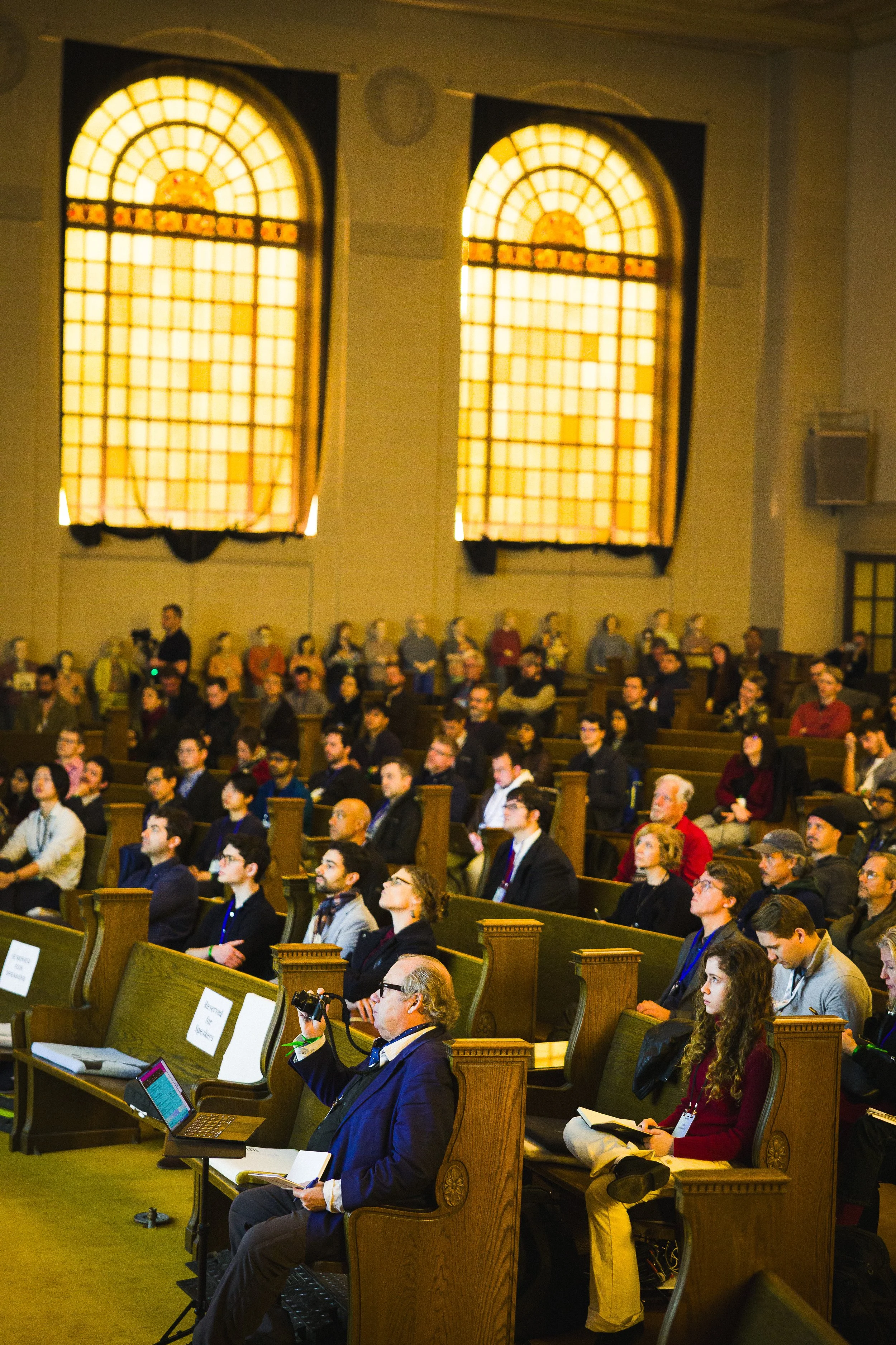 Audience attending a conference or presentation in a large room with stained glass windows.