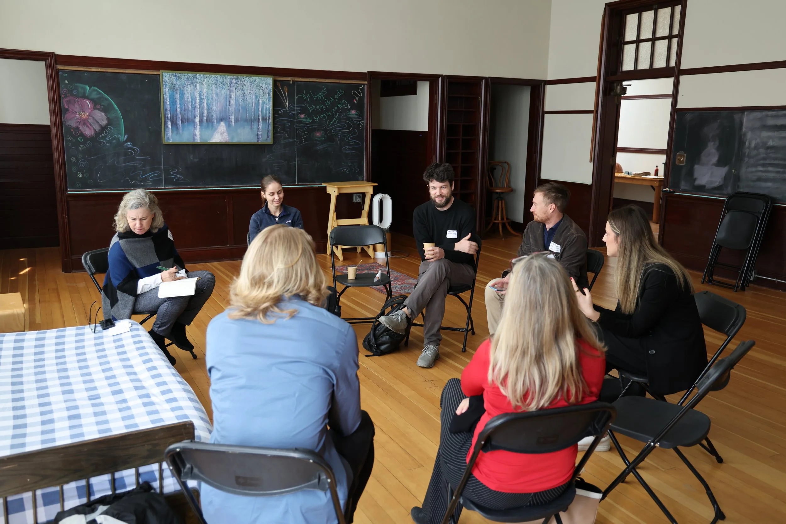 Group of people sitting in a circle in a classroom or meeting room with blackboards on the walls.