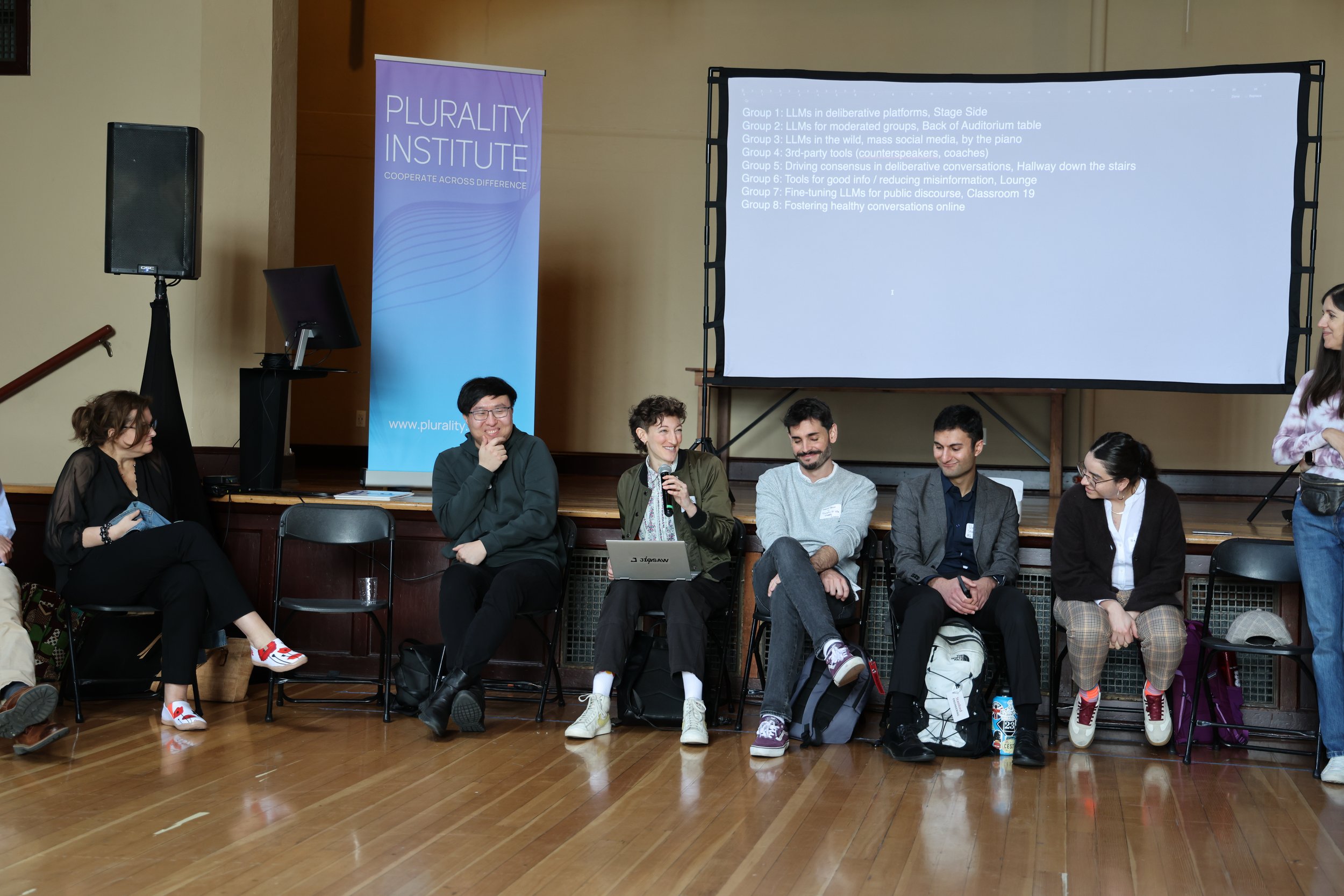 A group of seven people sitting on chairs in a panel discussion at an event. They are smiling and engaged, with a woman holding a microphone speaking. Behind them is a large screen displaying a list of discussion groups and a purple banner with the l
