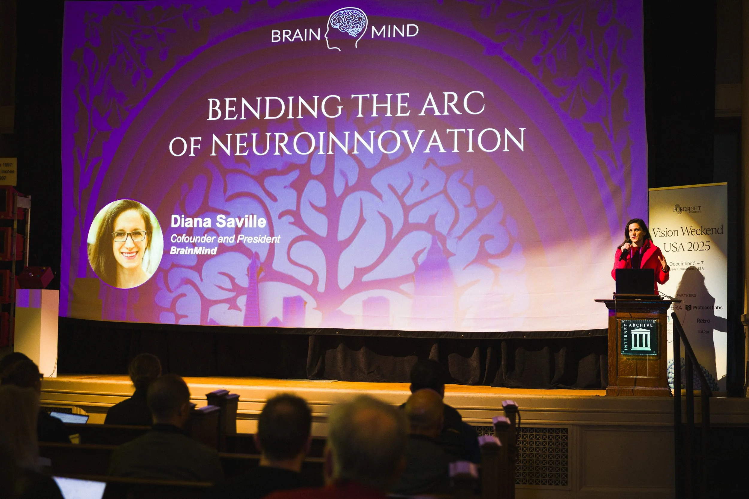 A woman in a red jacket is giving a presentation on stage with a large purple screen behind her. The screen displays the title 'Bending the Arc of Neuroinnovation' and a head silhouette logo with the words 'Brain' and 'Mind'. There is a photo of Dian