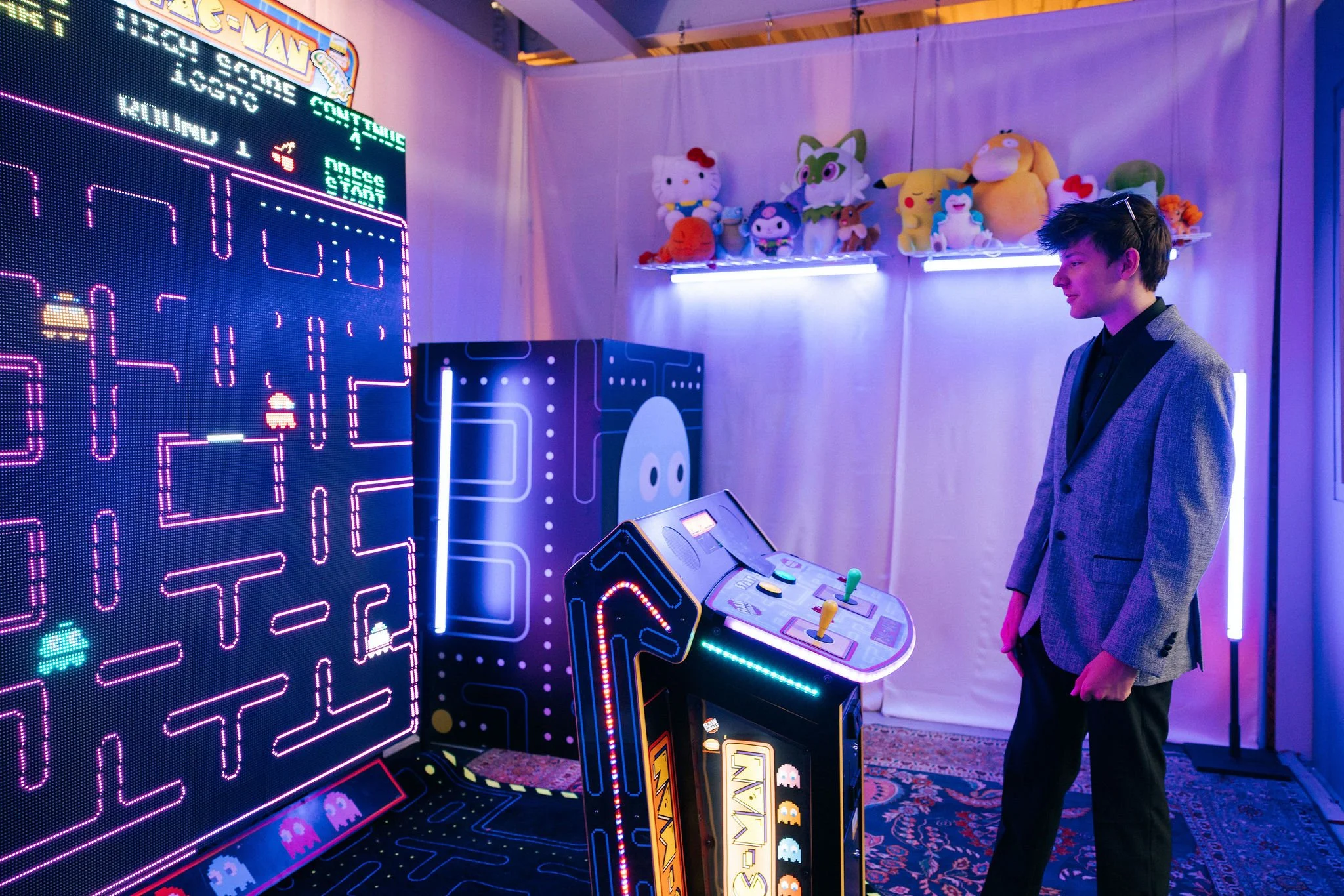 A young man wearing a gray blazer and black shirt stands in front of a Pac-Man arcade game with colorful neon lights. Behind him, stuffed toys are displayed on a shelf, including Hello Kitty and other characters. The room is lit with purple and blue 