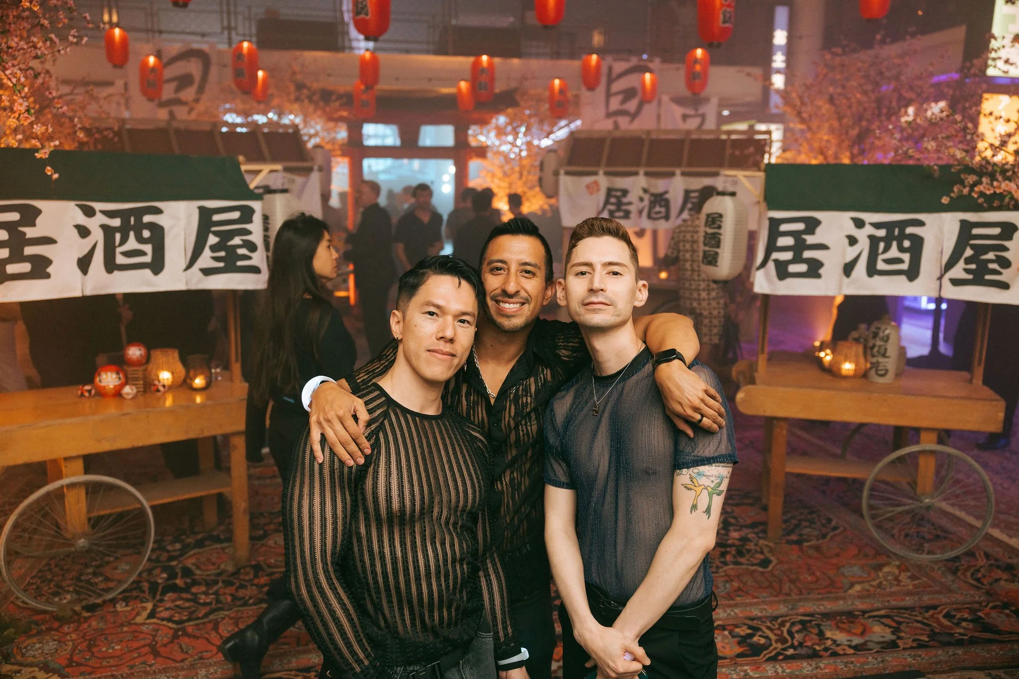 Three men posing together with arms around each other at an indoor party or celebration with Japanese lanterns and banners in the background.