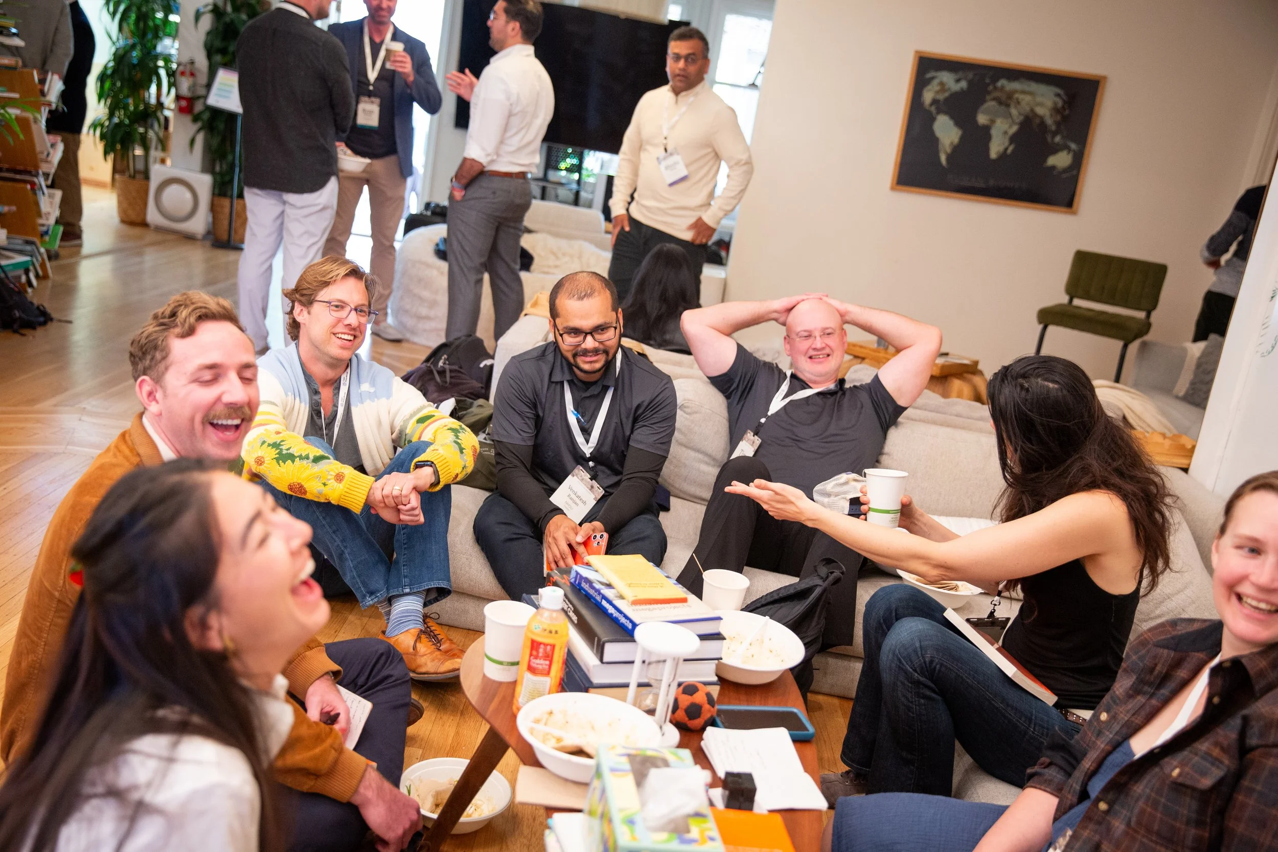 A group of smiling people, some sitting on a beige couch and others standing in a casual, social setting, with food and drinks on a coffee table.