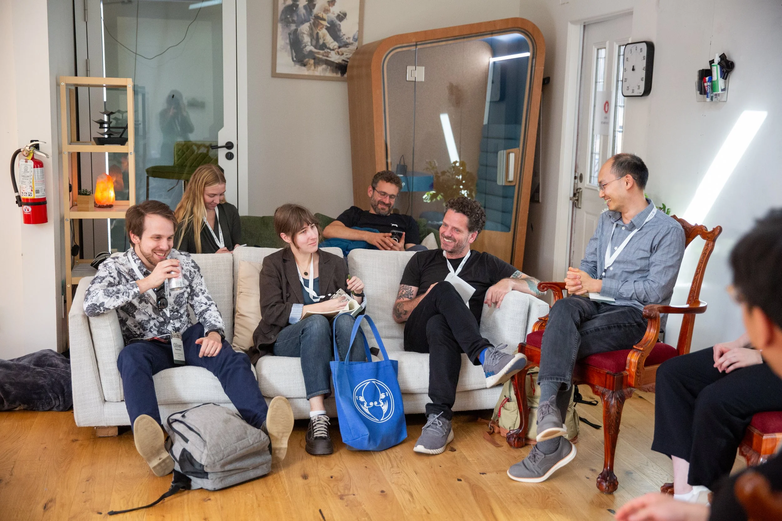 Group of seven people sitting and chatting in a cozy indoor space, with some holding drinks, smiling, and engaging in conversation.
