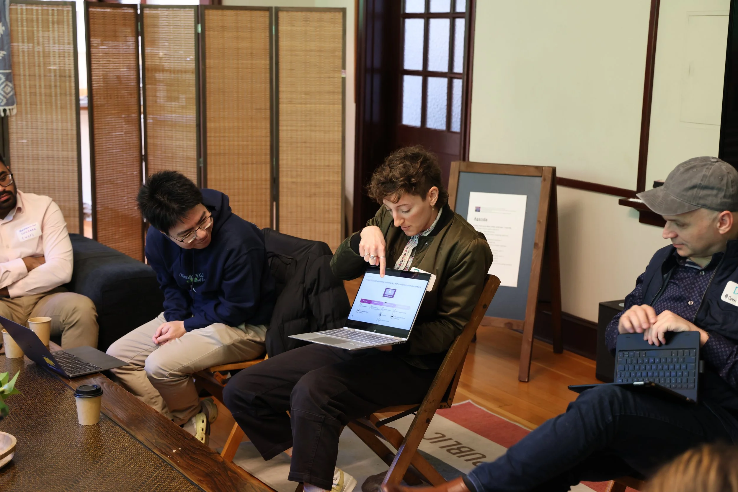 Four people seated around a table, one woman showing a laptop to a young man, with the others observing in a room with wooden floors and partitions.
