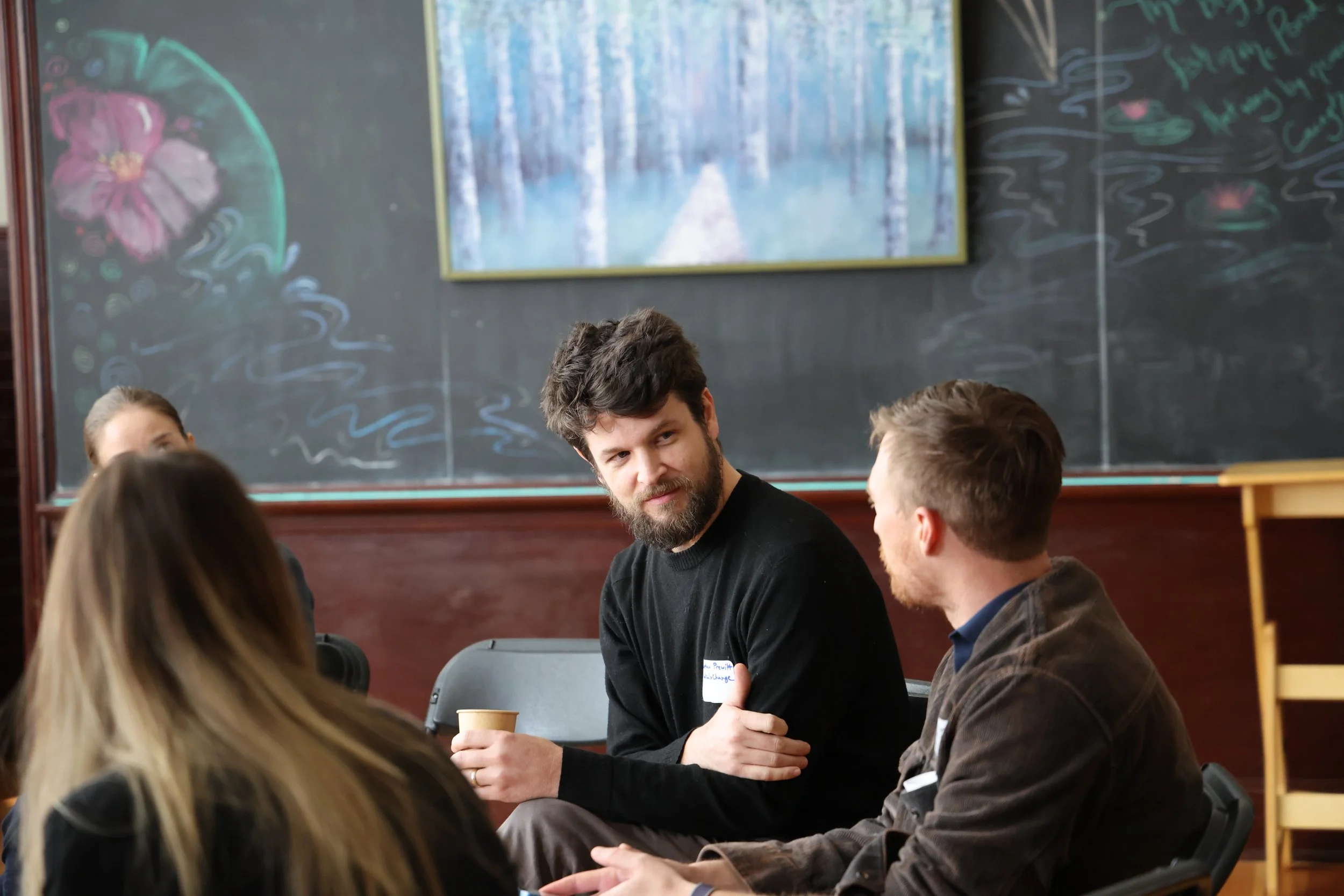 Group of people sitting and talking in a classroom with a chalkboard and artwork in the background.