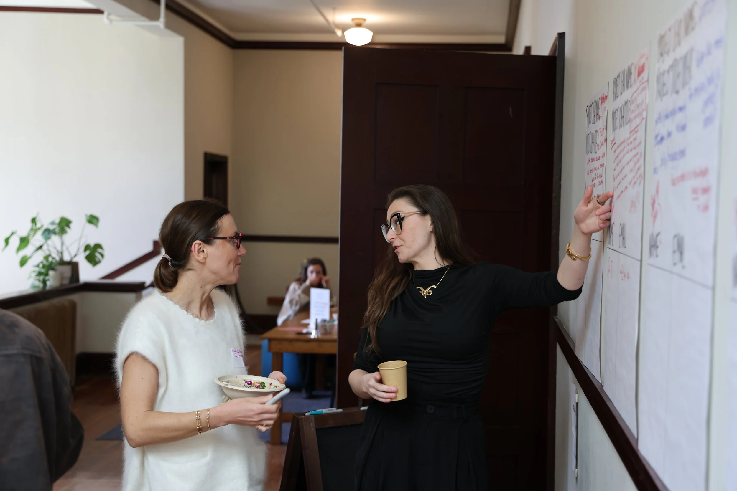 Two women in conversation near a whiteboard with notes, one gesturing towards the board, and a person in the background seated at a table.