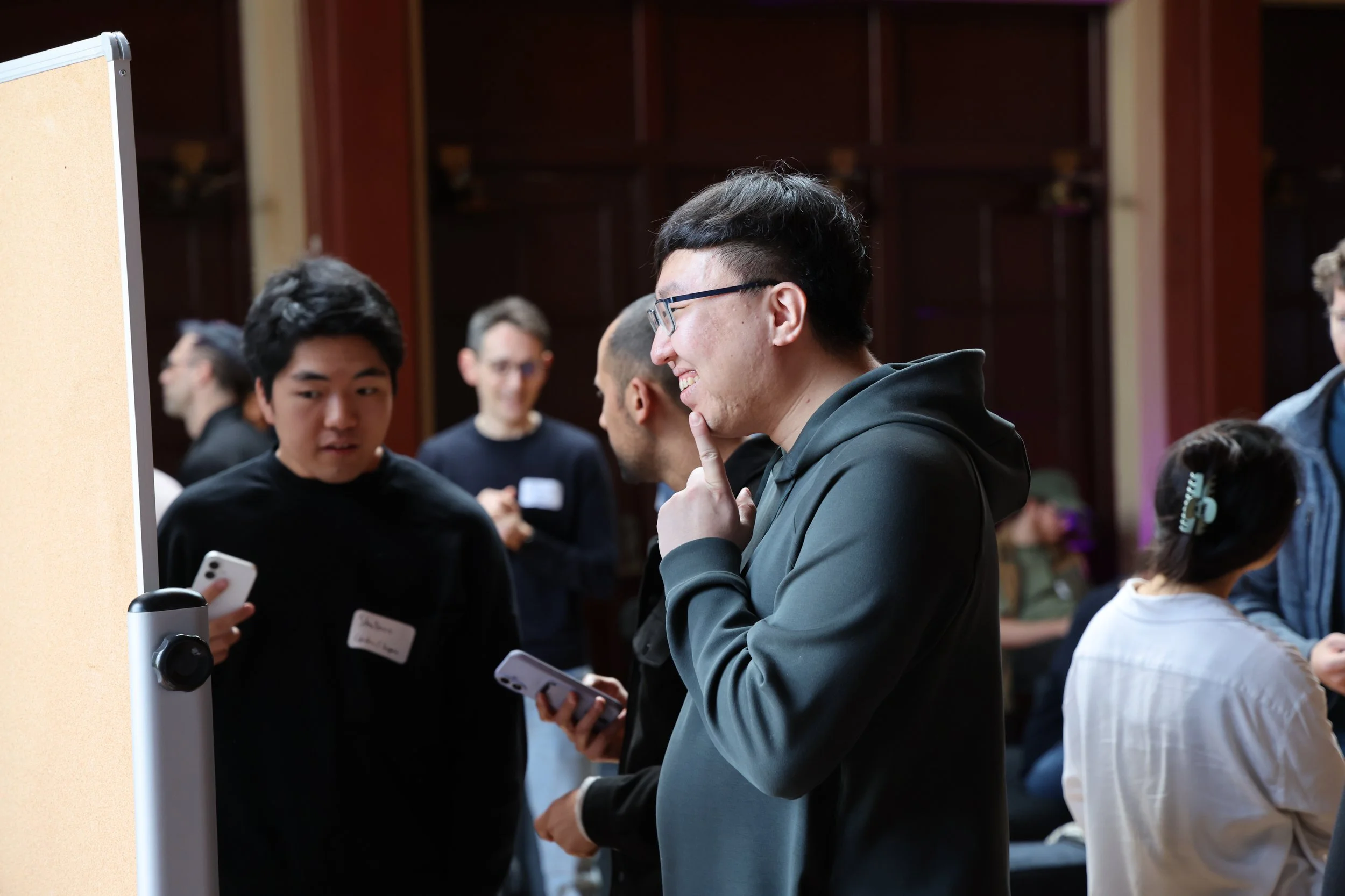 Group of people in a discussion at an indoor event, some holding smartphones, with a wooden wall in the background.