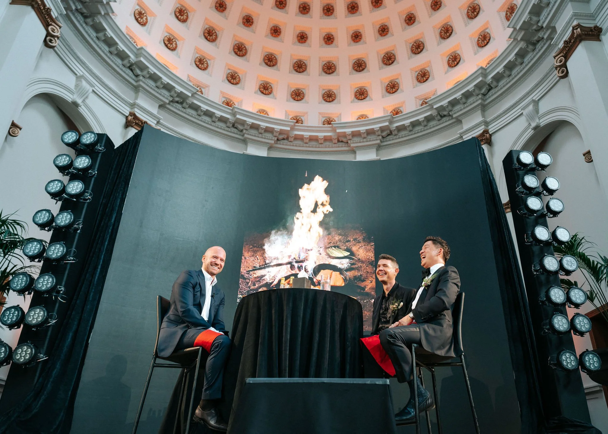 Three men in formal attire sitting around a table, laughing, with a fire display on a large screen behind them in a grand, domed hall.