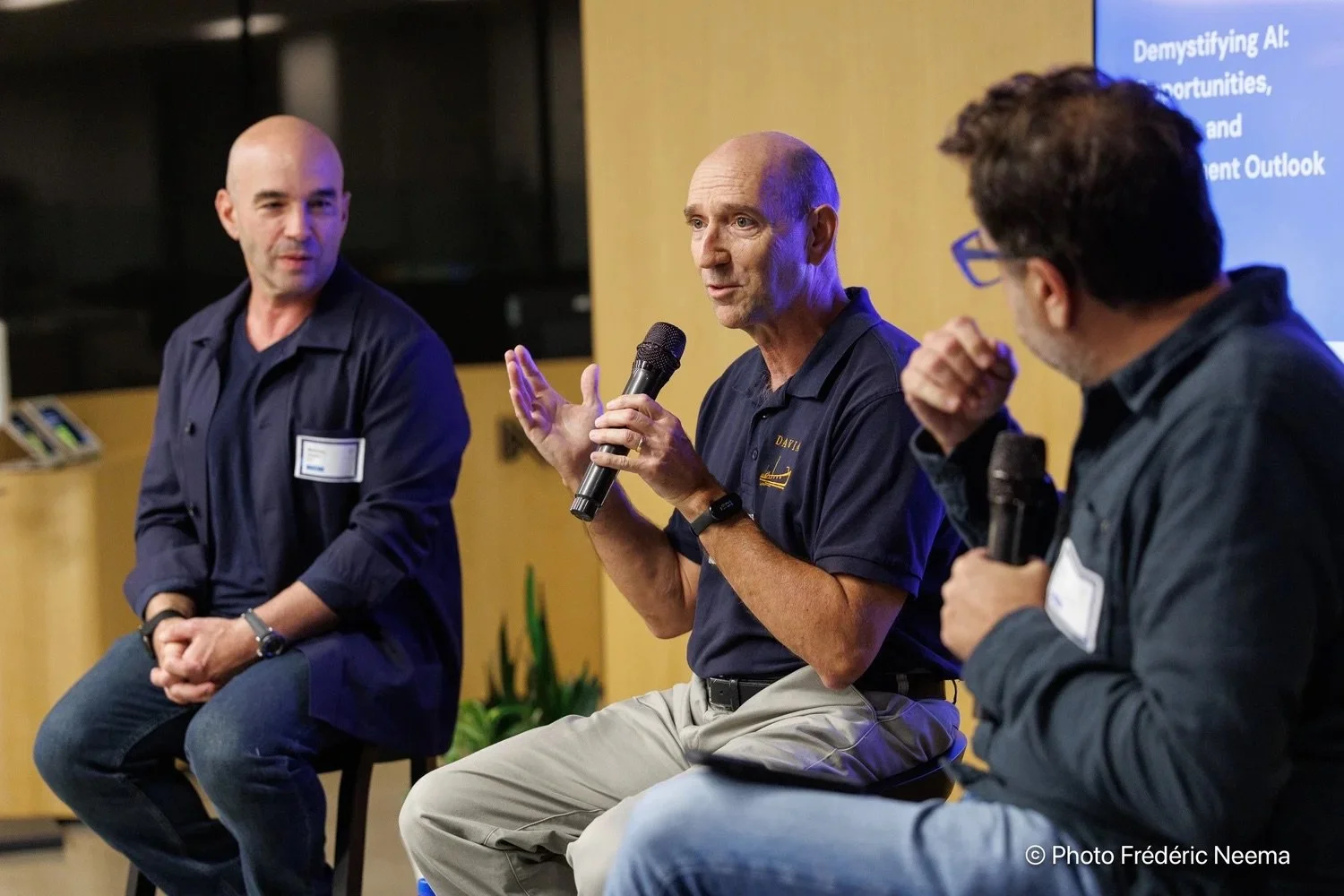 Three men participating in a panel discussion, with one man speaking into a microphone and the other two listening. They are indoors with a presentation screen in the background.