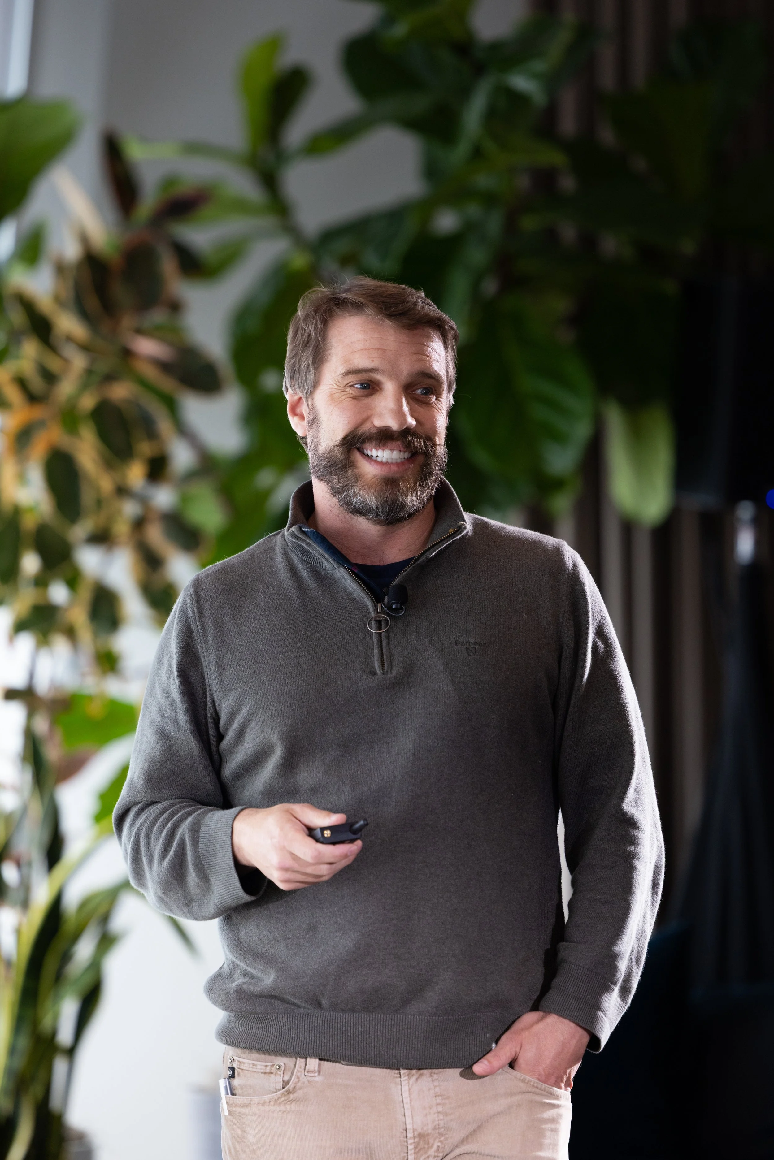 A man with a beard and mustache smiling, holding a remote control or small device, standing in front of green plants.