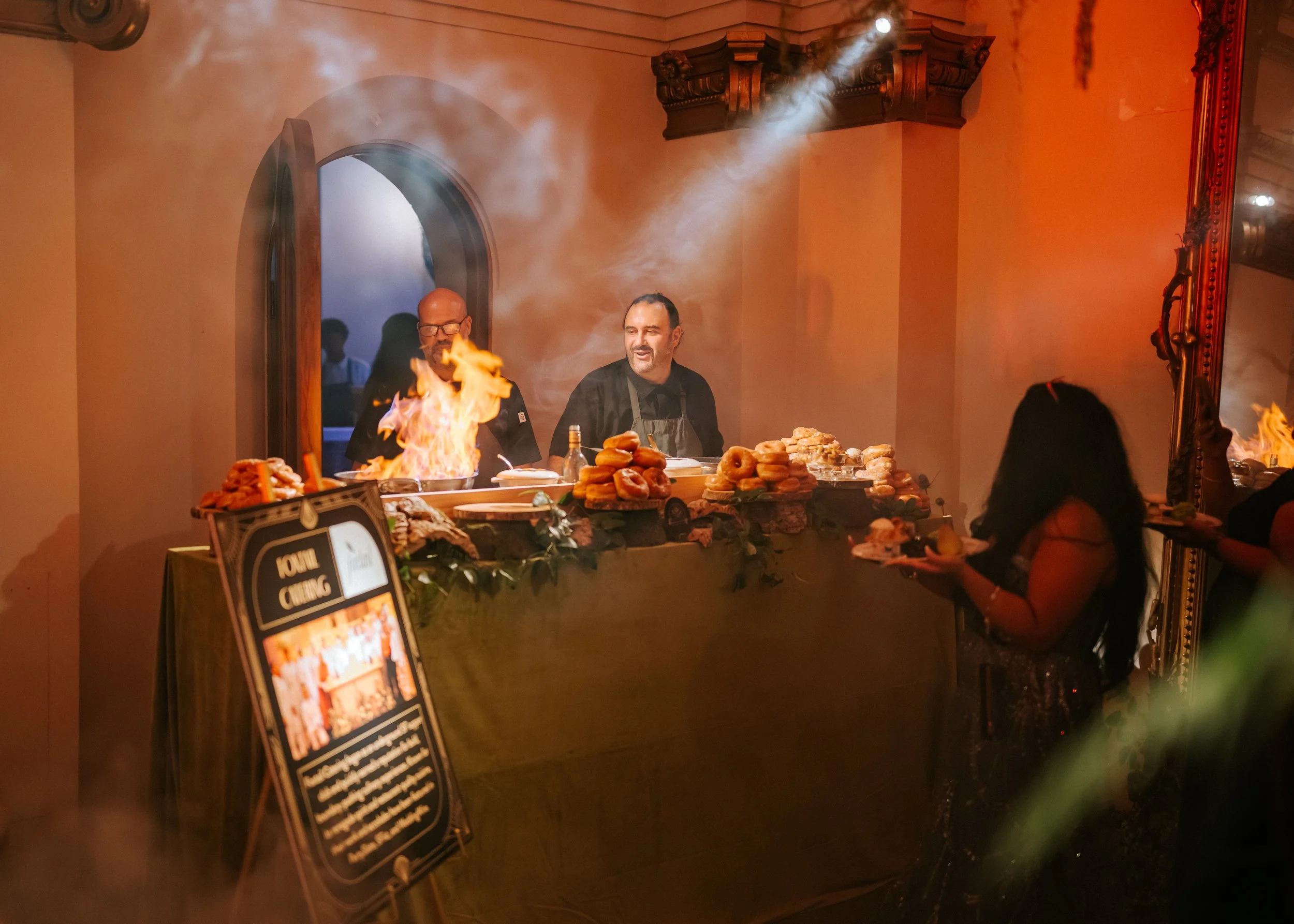 Chefs preparing food at a dinner event with flames on the table, surrounded by donuts and desserts, with a woman holding a plate in the foreground and a signboard in the corner.