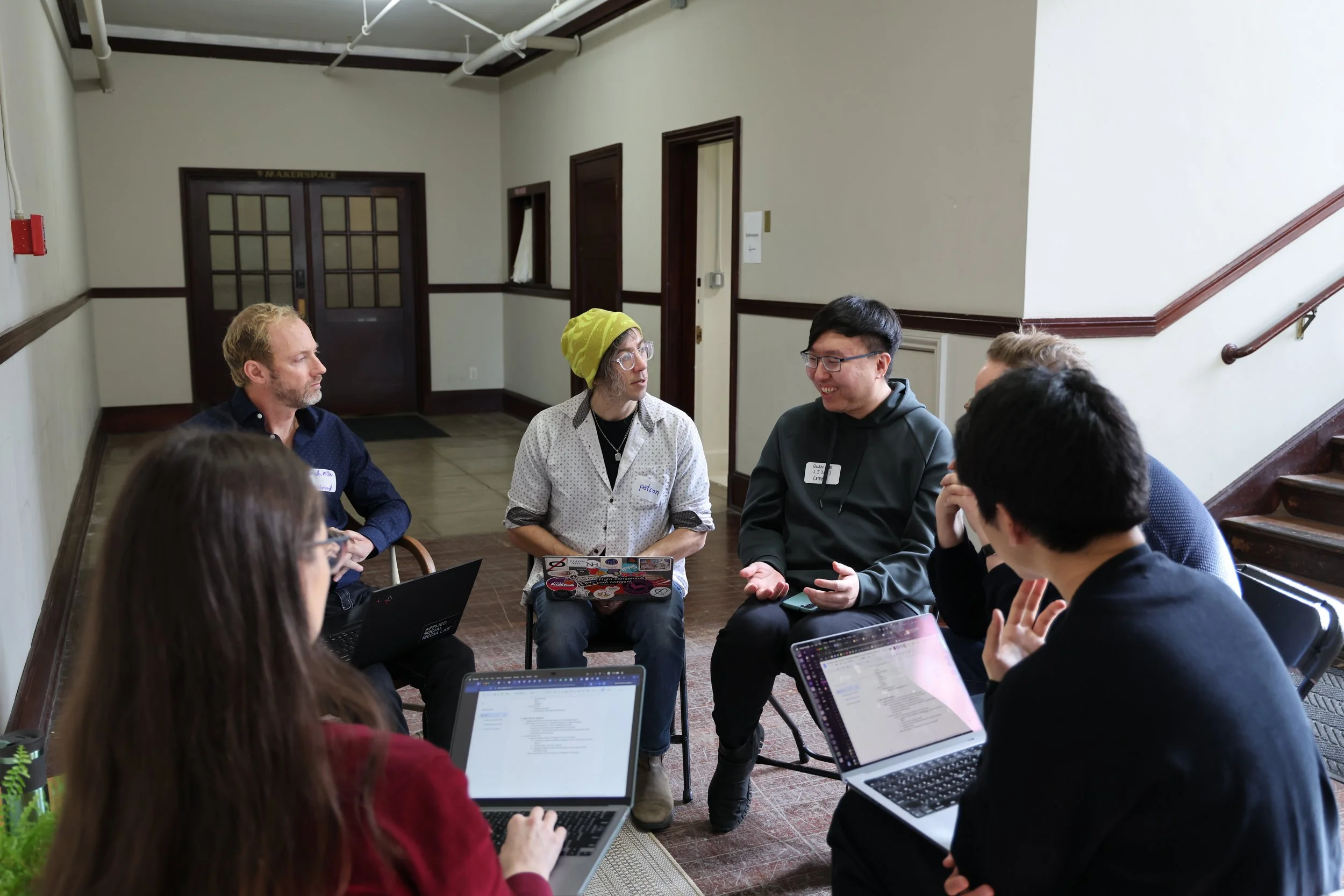 Group of six people sitting in a circle having a discussion, some with laptops open.