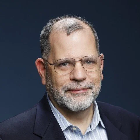 A middle-aged man with glasses, a beard, and short gray hair, wearing a suit and a light blue shirt, smiling against a dark background.