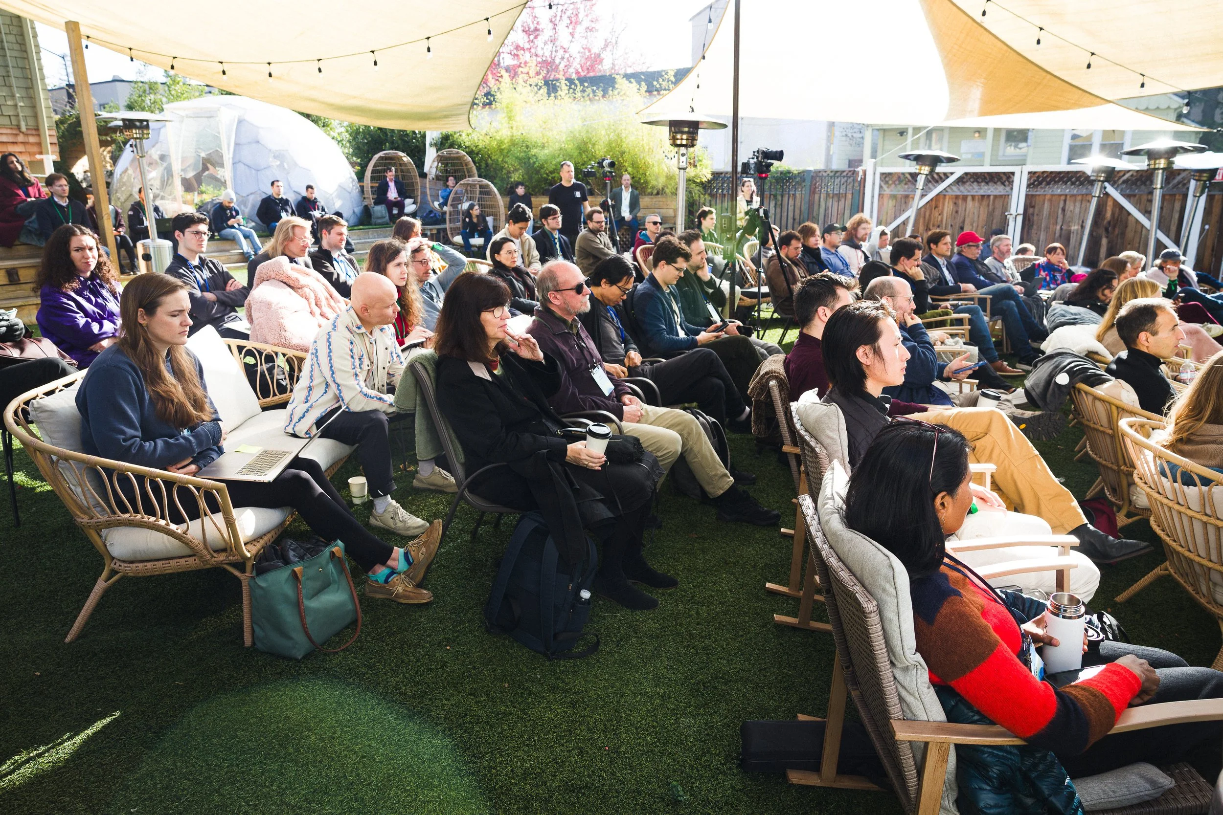 An outdoor conference or seminar with an audience seated under a canopy, with string lights overhead. Attendees are seated in various chairs on a grassy area, paying attention to a speaker or presentation.
