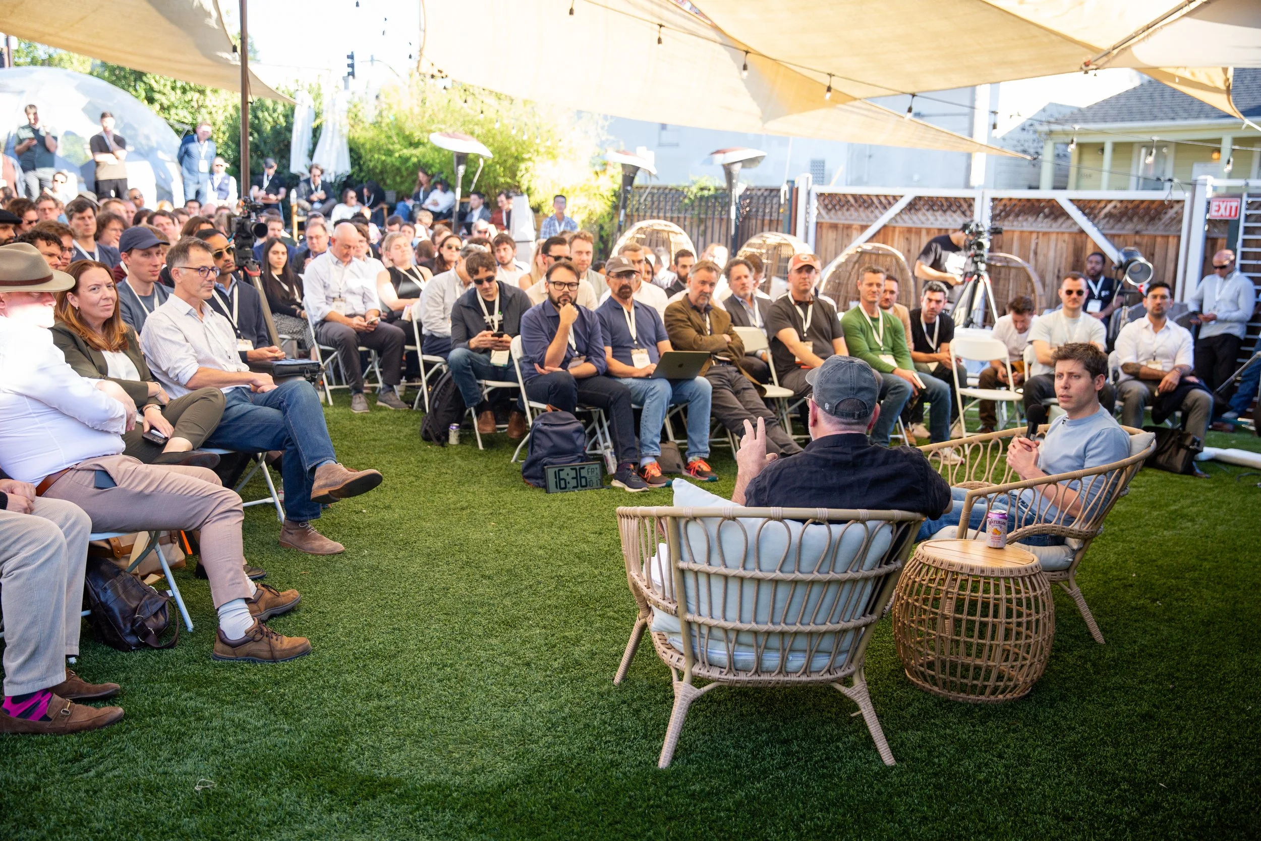 People attending an outdoor event under a large beige canopy, seated on chairs and lounge furniture, listening to speakers at a panel. The setting has green grass and a fence in the background.