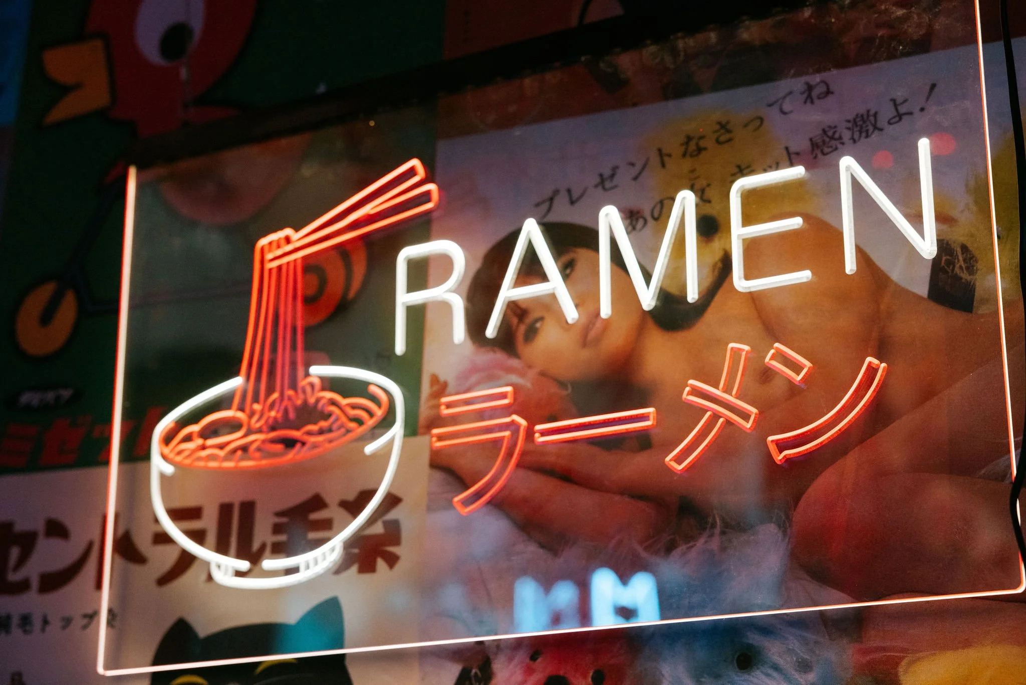 Neon sign with Japanese characters and the word 'RAMEN' in English, featuring an illustration of a bowl of ramen noodles with chopsticks. Behind the sign, there is a poster with a woman reclining on a fluffy surface.