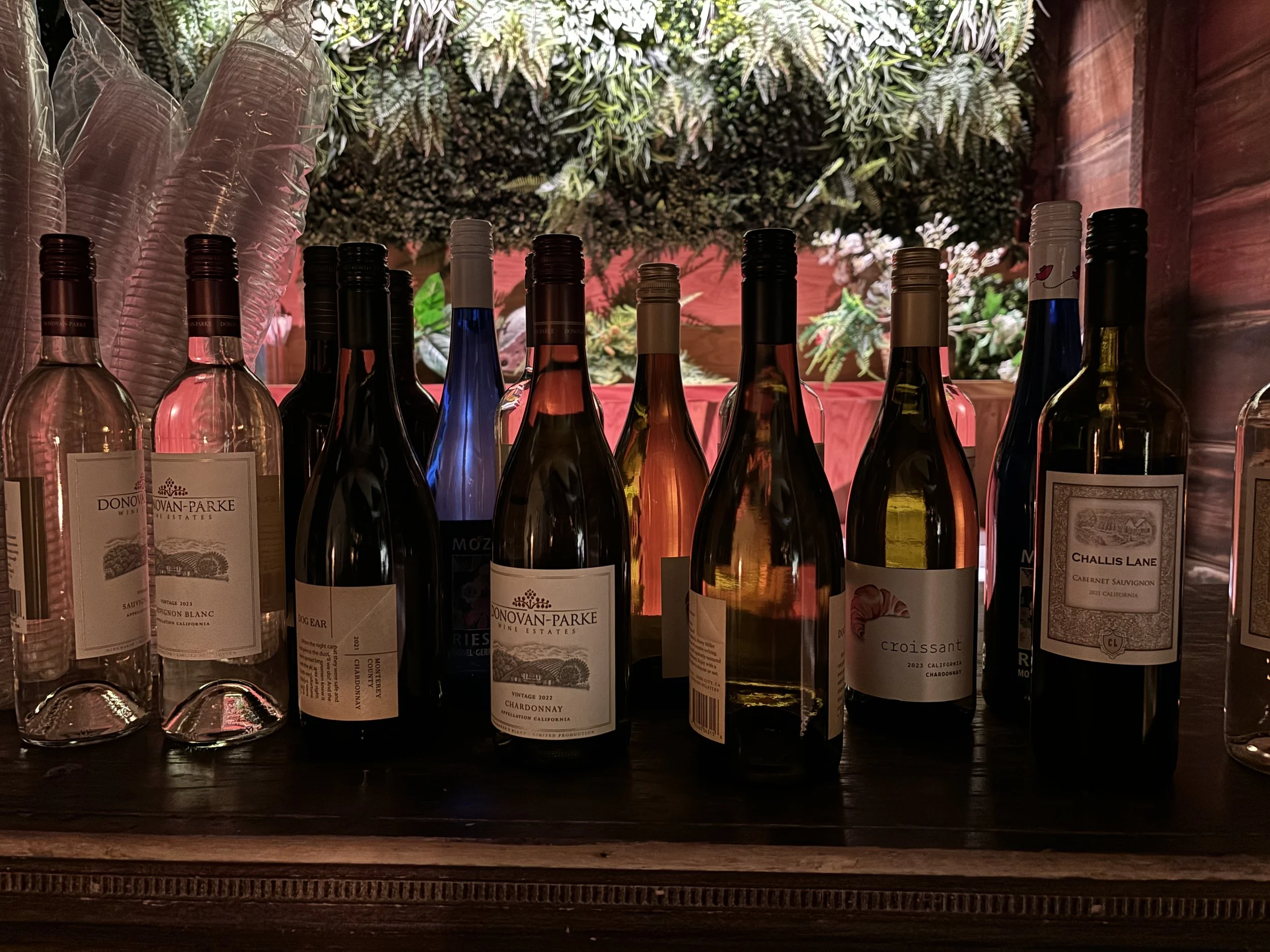 Various bottles of wine, including Chardonnay, Cabernet Sauvignon, and others, arranged on a wooden shelf with decorative greenery and flowers in the background.